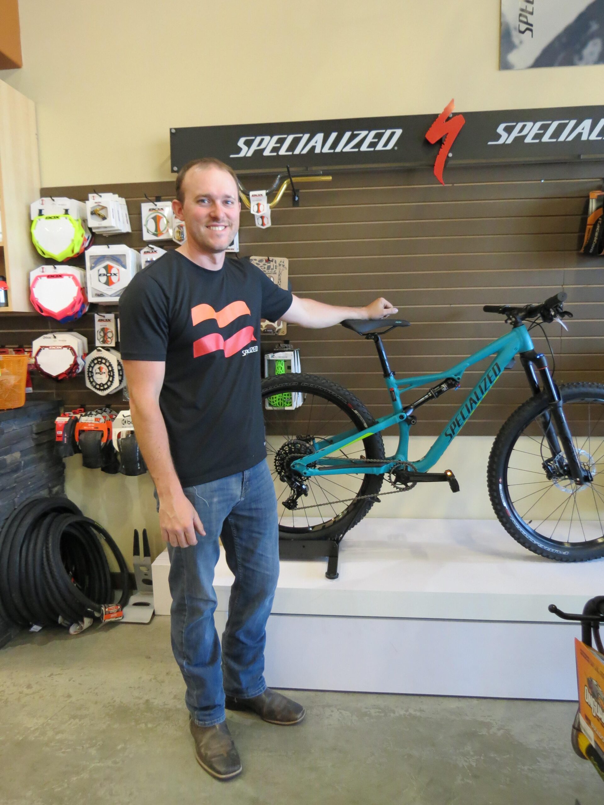 A man standing next to a teal mountain bike in a bike shop. He is smiling and wearing a black t-shirt with a logo, paired with blue jeans and brown boots. The store features various cycling products displayed on the walls, including accessories and bike tires.
