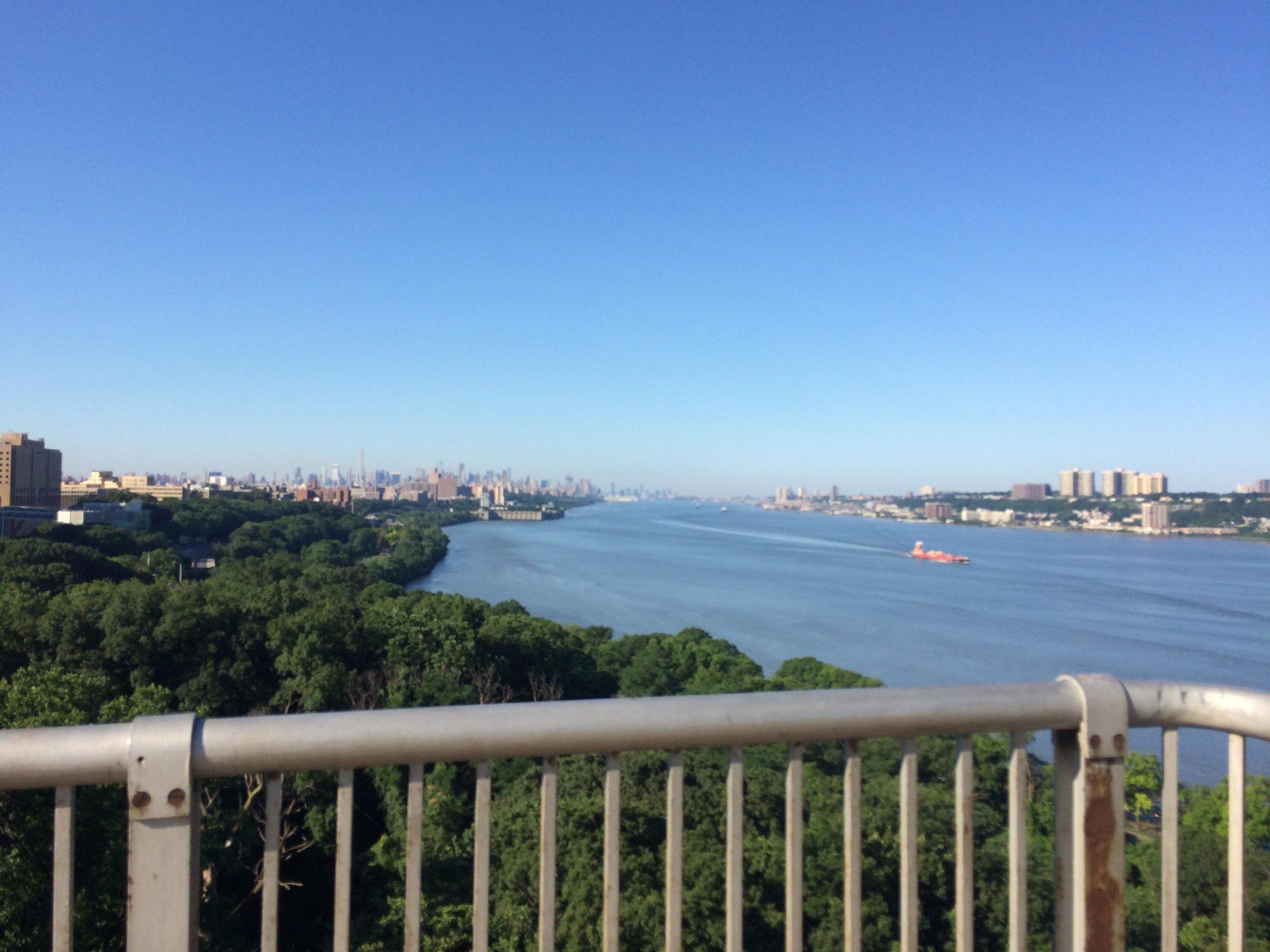 A panoramic view of a river bordered by lush greenery, with a distant city skyline featuring tall buildings under a clear blue sky. A boat is seen traveling along the waterway in the foreground. West Street Greenway mountain bike trail.