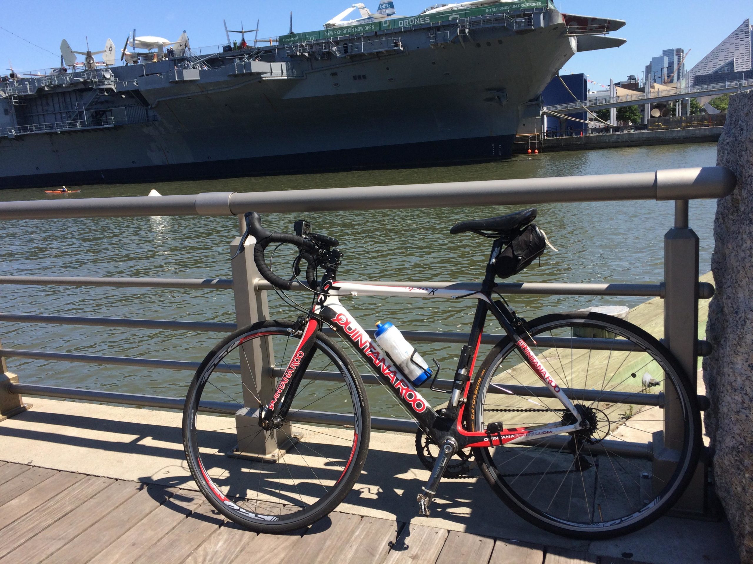 A black and red road bicycle parked by a waterfront railing, with a large military ship in the background and city buildings visible on the horizon. The scene is set on a sunny day, with calm water and a few people in kayaks nearby. West Street Greenway mountain bike trail.