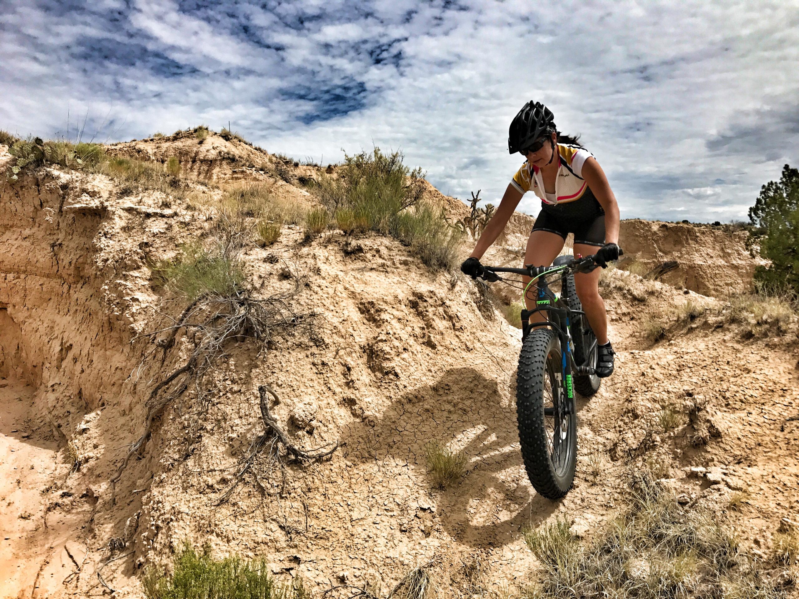 A person riding a mountain bike on a rugged dirt trail surrounded by dry terrain and sparse vegetation, with a cloudy sky overhead. The cyclist is wearing a helmet and sports attire, focusing on maneuvering the rocky landscape. Mariposa Fat Bike Trails mountain bike trail.