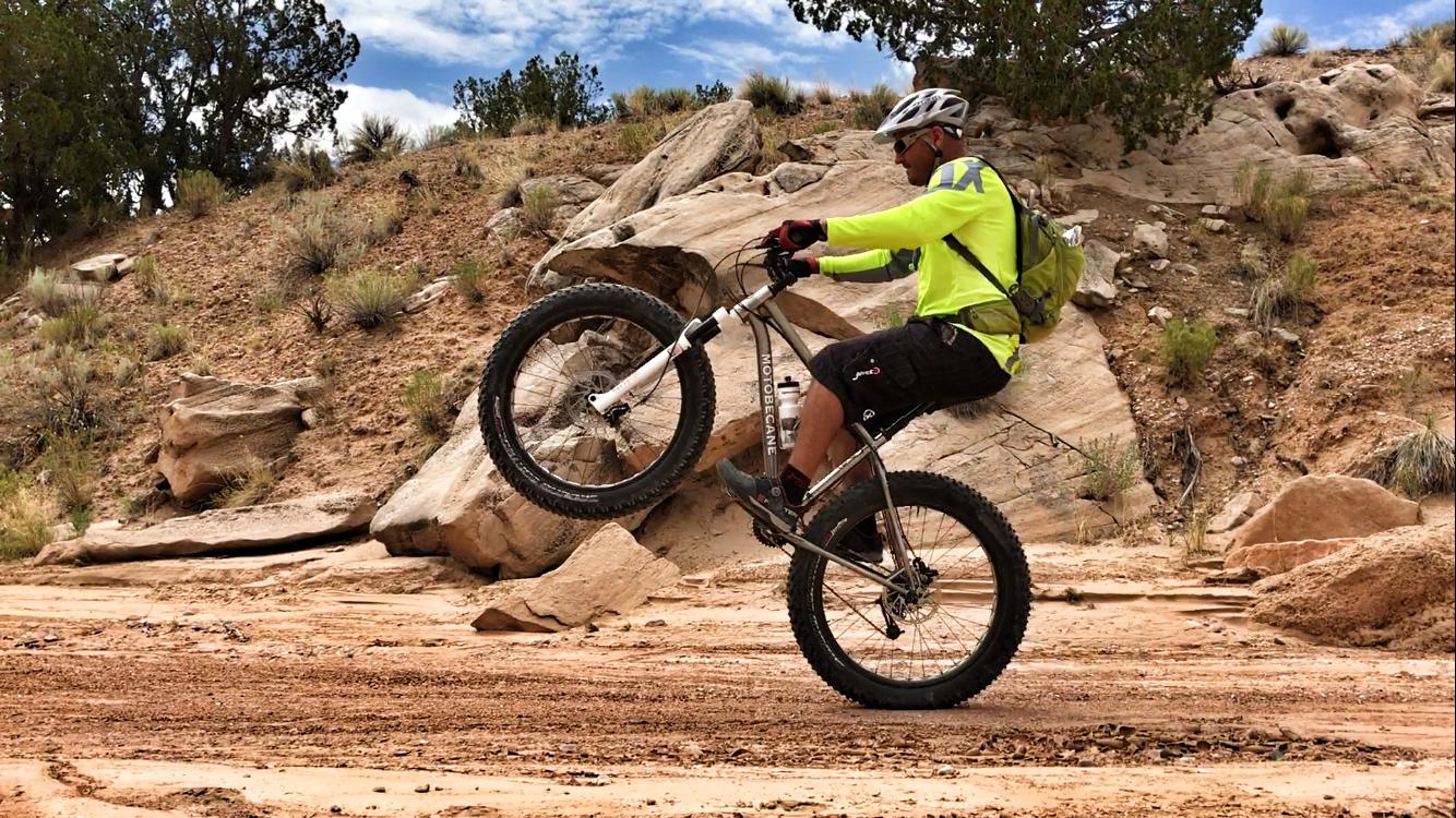 A cyclist wearing a bright yellow jersey and helmet performs a wheelie on a mountain bike on a rocky dirt path, surrounded by sparse vegetation and rocky terrain under a blue sky. Mariposa Fat Bike Trails mountain bike trail.