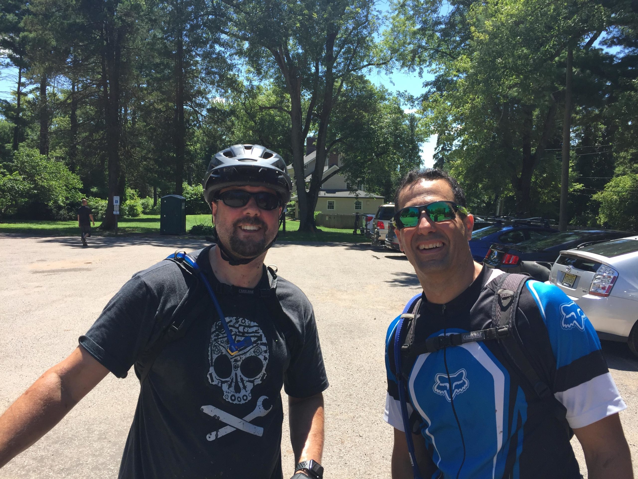Two men smiling at the camera, wearing bicycle helmets and sunglasses. One man has a casual black shirt with a skull design, while the other wears a blue and white athletic biking jersey. They are outdoors in a sunny area with trees in the background and cars parked nearby. Six Mile Run mountain bike trail.
