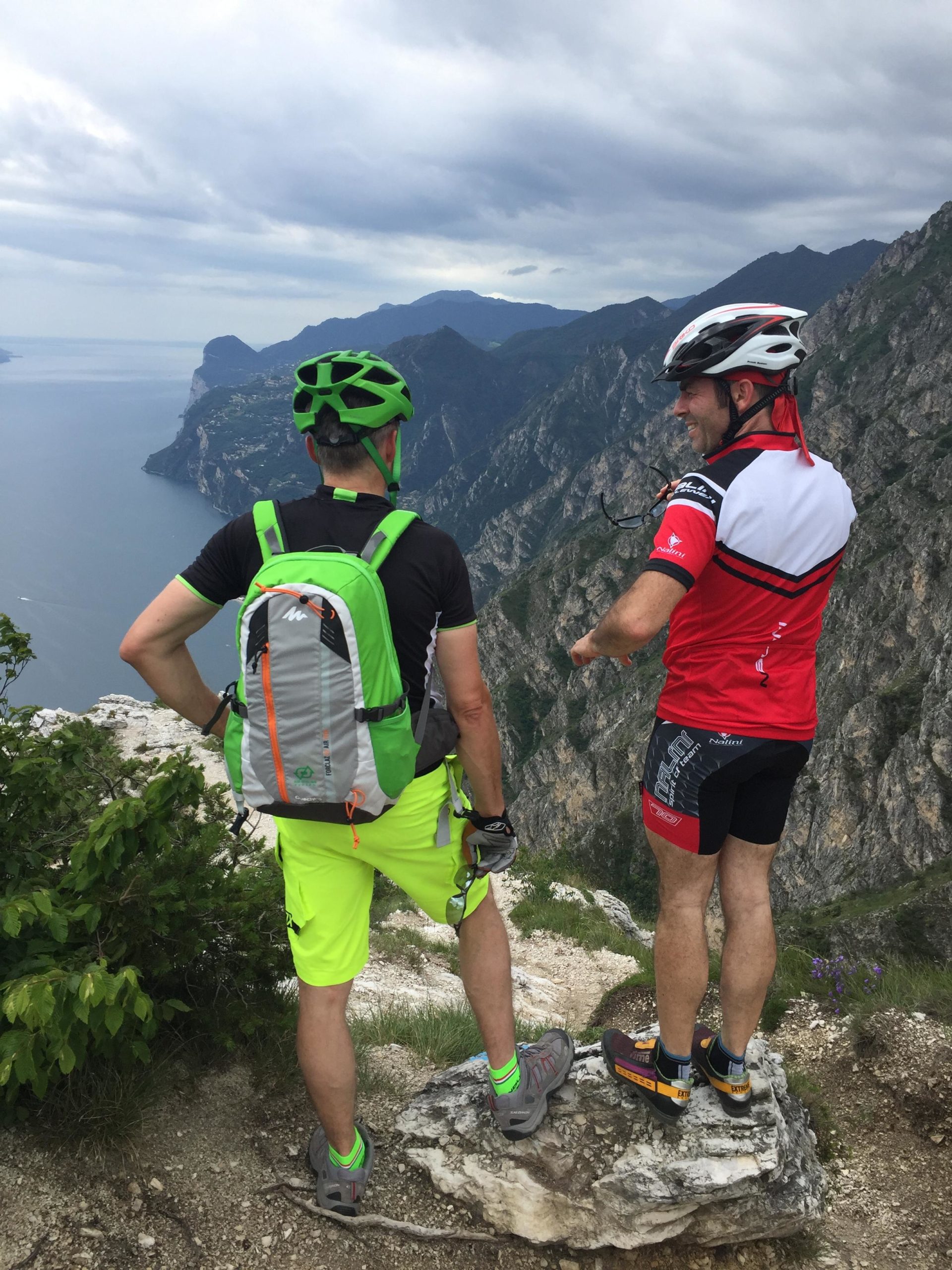 Two cyclists, one in a bright green helmet and backpack and the other in a red jersey, stand on a rocky ledge overlooking a scenic view of mountains and a lake. They appear to be discussing the landscape, with cloudy skies overhead and greenery in the foreground. Vesio Tremalzo mountain bike trail.