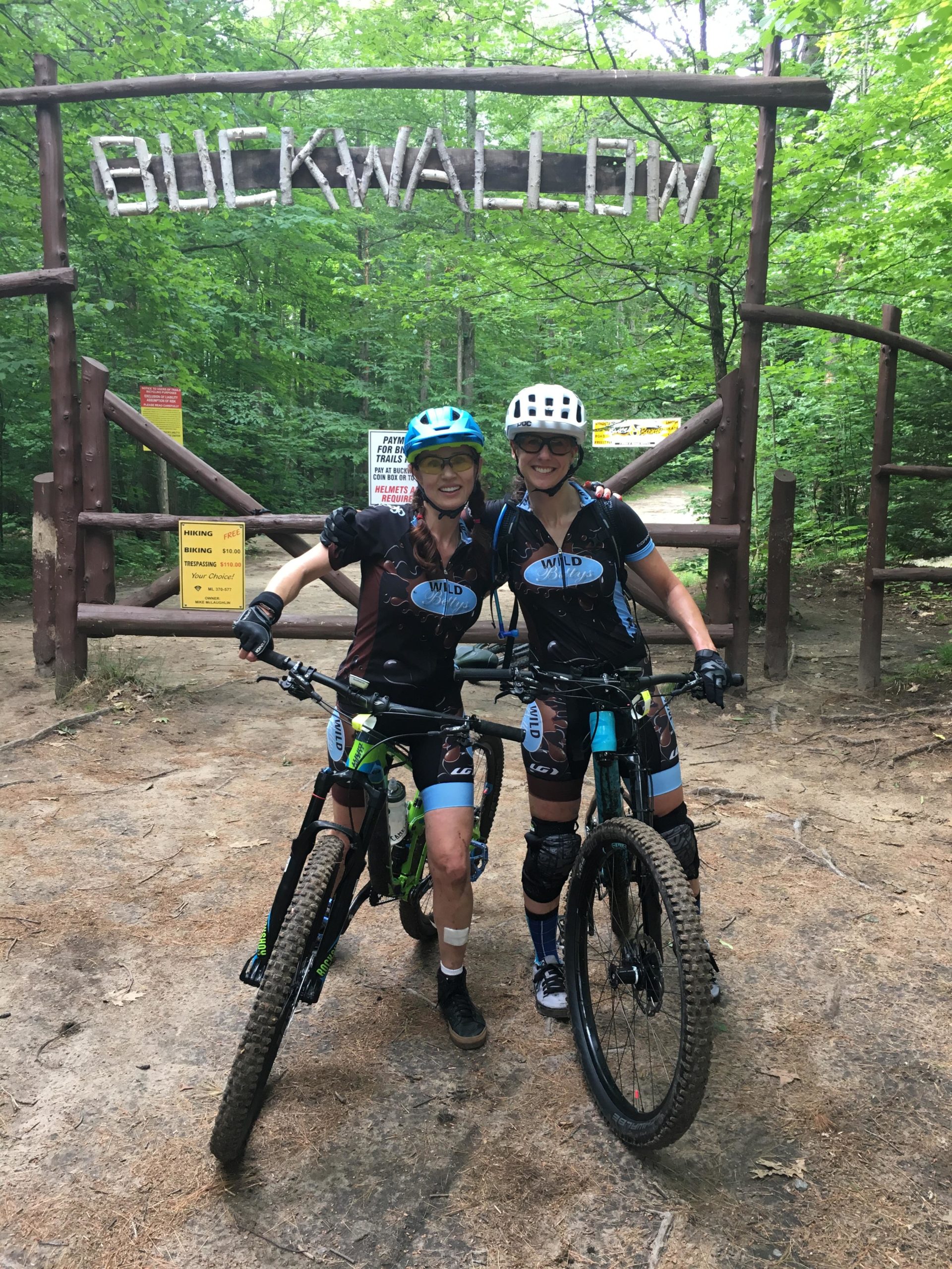 Two cyclists posing in front of a wooden sign that reads "BUCKWALLOW." They are wearing cycling gear and helmets, with bikes parked beside them in a wooded area. The background features green trees, and signs for biking and hiking can be seen nearby. Buckwallow mountain bike trail.
