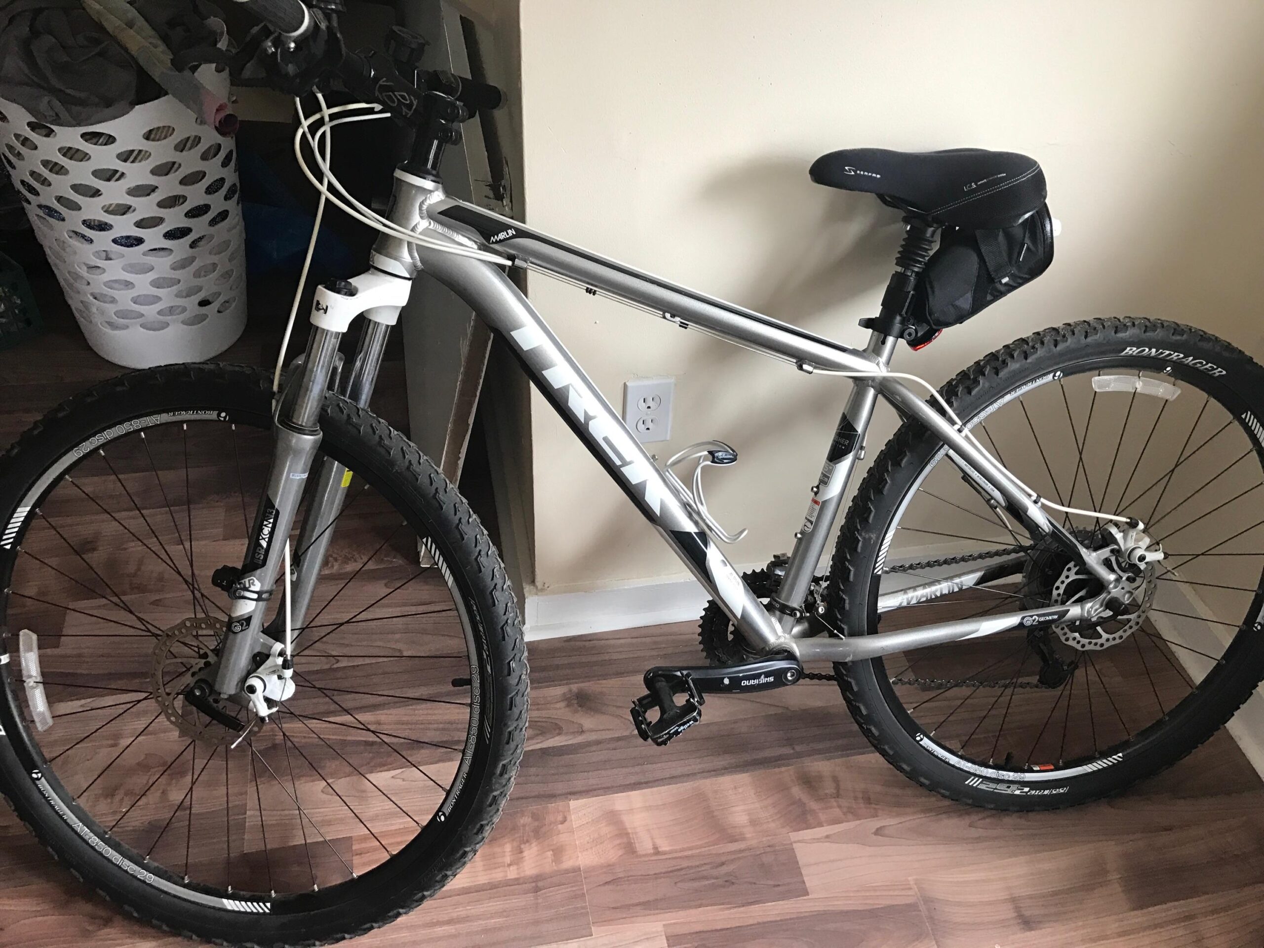 Trek Marlin: A silver mountain bike with a black seat and small bag attached, positioned indoors on a wooden floor. The bike features thick tires with prominent tread, disc brakes, and visible gear components. In the background, there's a laundry basket and a doorway.