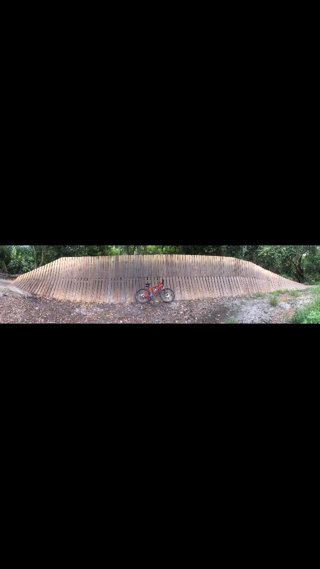 A mountain bike is leaned against a wooden pump track surrounded by trees. The track features multiple sloped, wooden walls designed for cycling, with a dirt path running alongside it. The scene captures a natural outdoor setting ideal for biking activities. Markham Park mountain bike trail.