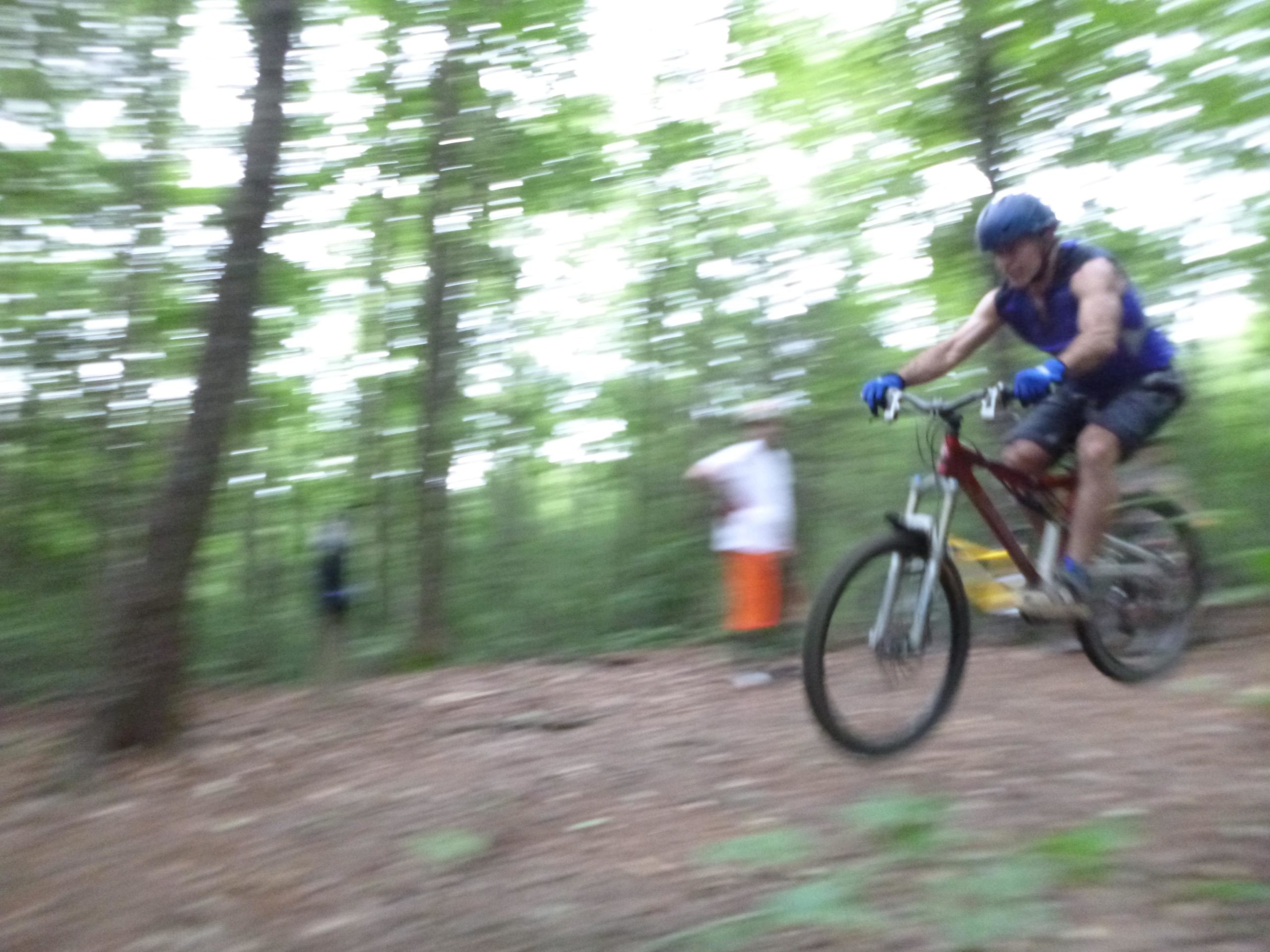 A mountain biker in a blue tank top, helmet, and gloves rides swiftly through a wooded area, captured in motion with a blurred background of trees. A second person is faintly visible in the background. Dark Mountain Trail mountain bike trail.