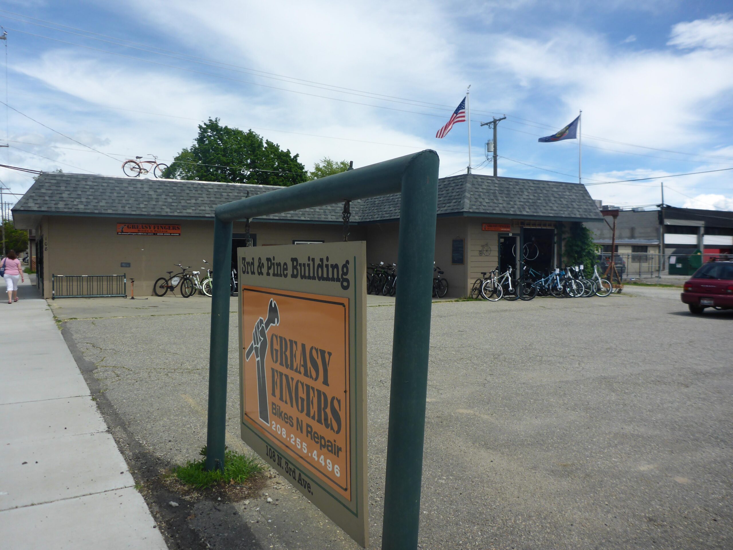 A view of the exterior of a bike repair shop named "Greasy Fingers," located in the 3rd & Pine Building. The shop features bicycles displayed outside and has signage prominently stating its name. The scene includes an American flag and another flag flying on the roof. A pedestrian walks along the sidewalk, and a parked car is visible in the foreground. The sky is partly cloudy.