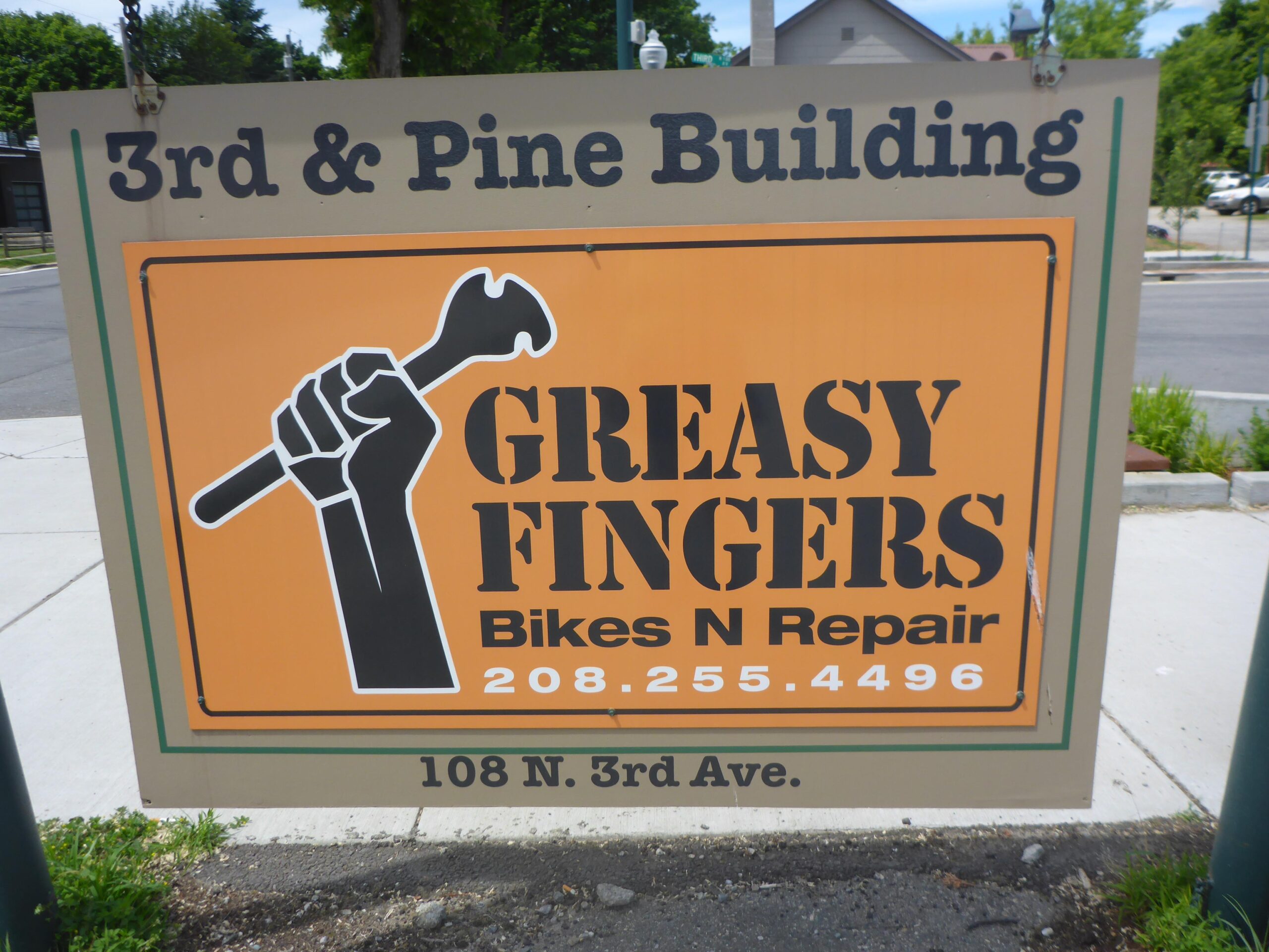 Sign for "Greasy Fingers Bikes N Repair" located at 108 N. 3rd Ave., featuring a graphic of a raised fist holding a wrench against an orange background. The top of the sign reads "3rd & Pine Building." Contact number displayed is 208.255.4496.