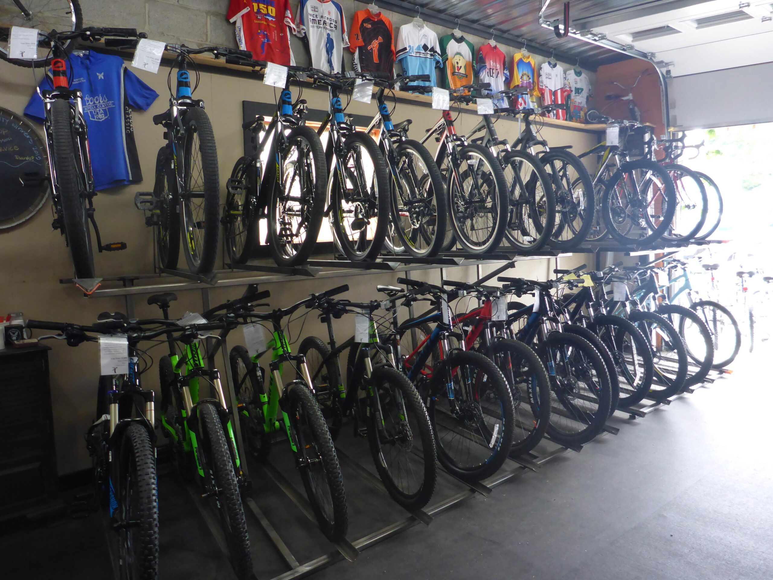 A display of various mountain bikes arranged on shelves in a bike shop, with colorful cycling jerseys hanging above. The bikes feature different colors and styles, indicating a range of options available for customers.