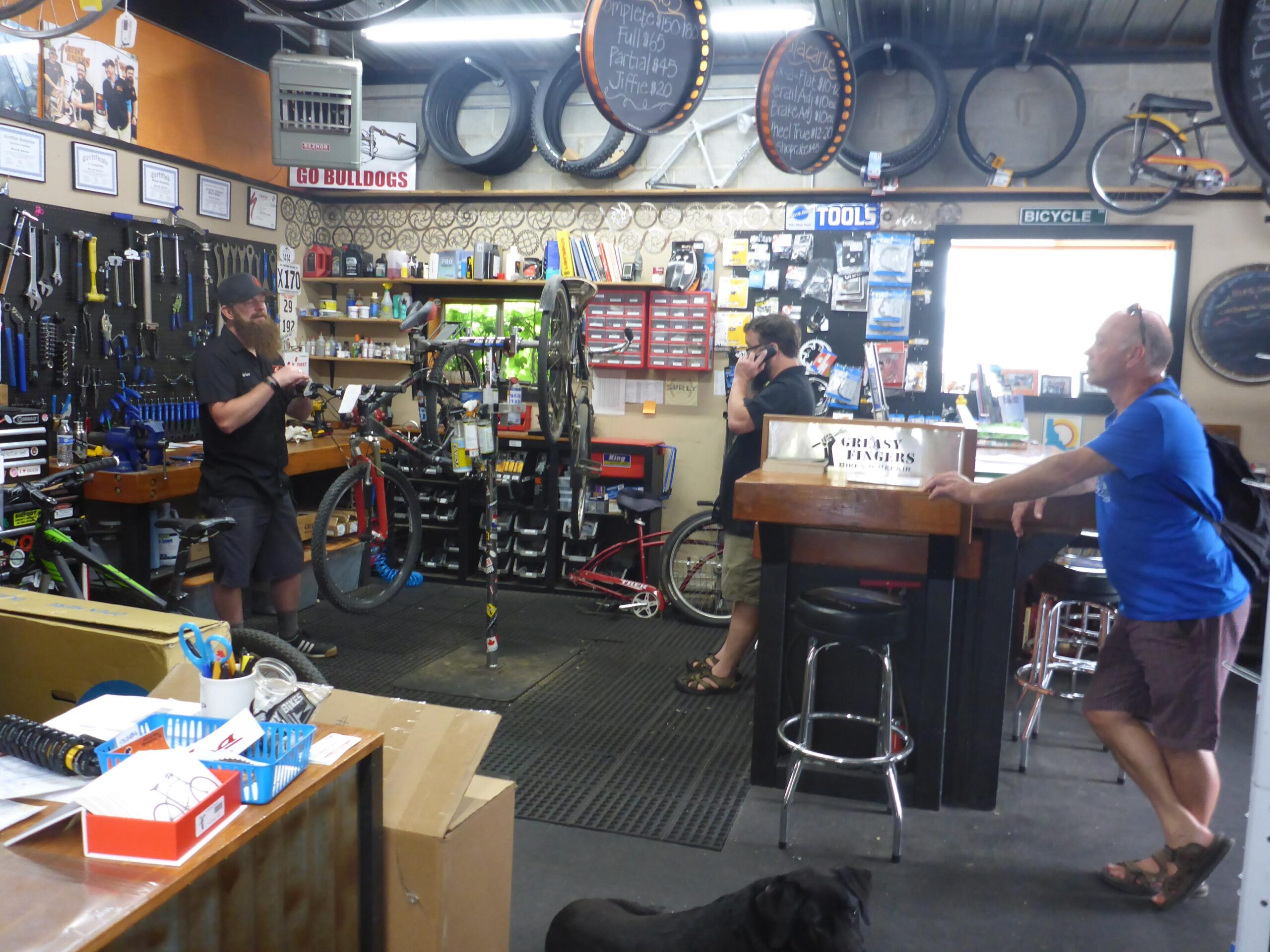 A busy bicycle shop interior featuring a man with a long beard examining a bike while standing next to a repair stand. Another man in a dark shirt is talking on the phone, and a third man in a blue shirt is leaning against the counter. Various bike tools and parts are visible on the walls, with bicycles and tires hanging above. In the foreground, there's a black dog resting on the floor.