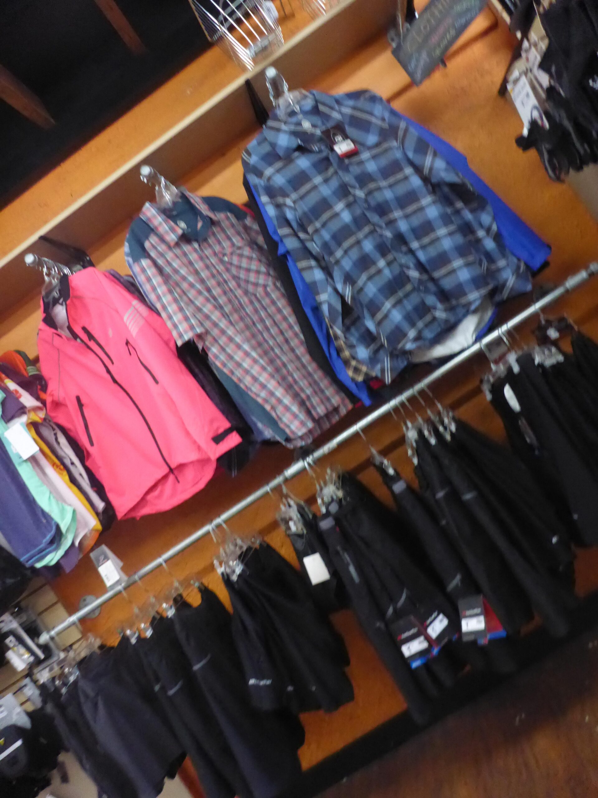 Display of clothing items in a retail store, featuring various shirts in colors like pink and blue, and a selection of black pants hanging on racks. The shirts include plaid and solid designs, arranged on hangers against an orange wall.
