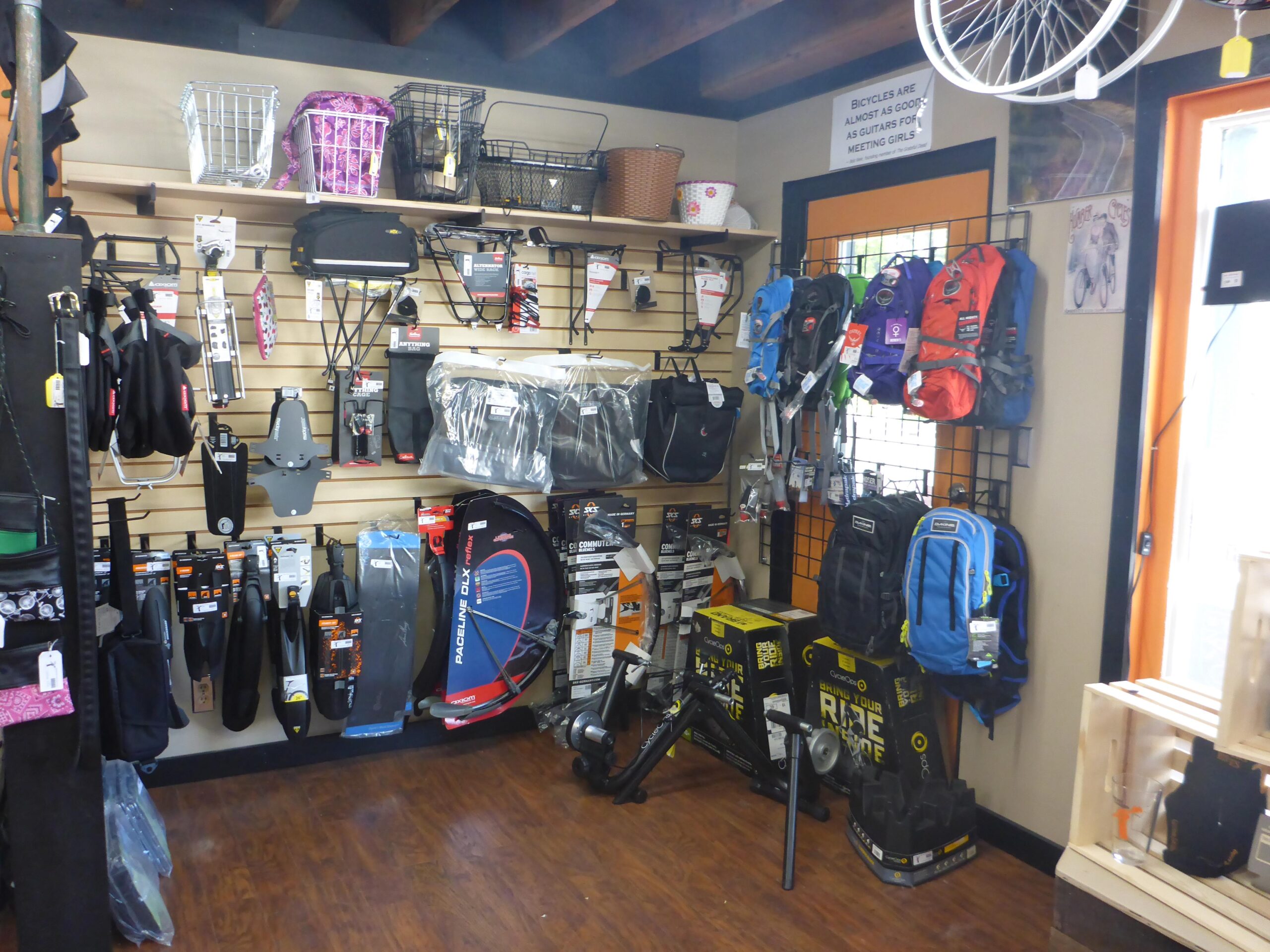 A display area in a bike shop featuring various cycling accessories such as saddlebags, backpacks, fenders, and tire maintenance tools. The wall is lined with organized shelves and hooks holding different items, and there&rsquo;s a bicycle wheel hanging from the ceiling. A humorous sign about bicycles is visible on the wall, with wooden flooring completing the cozy shop atmosphere.
