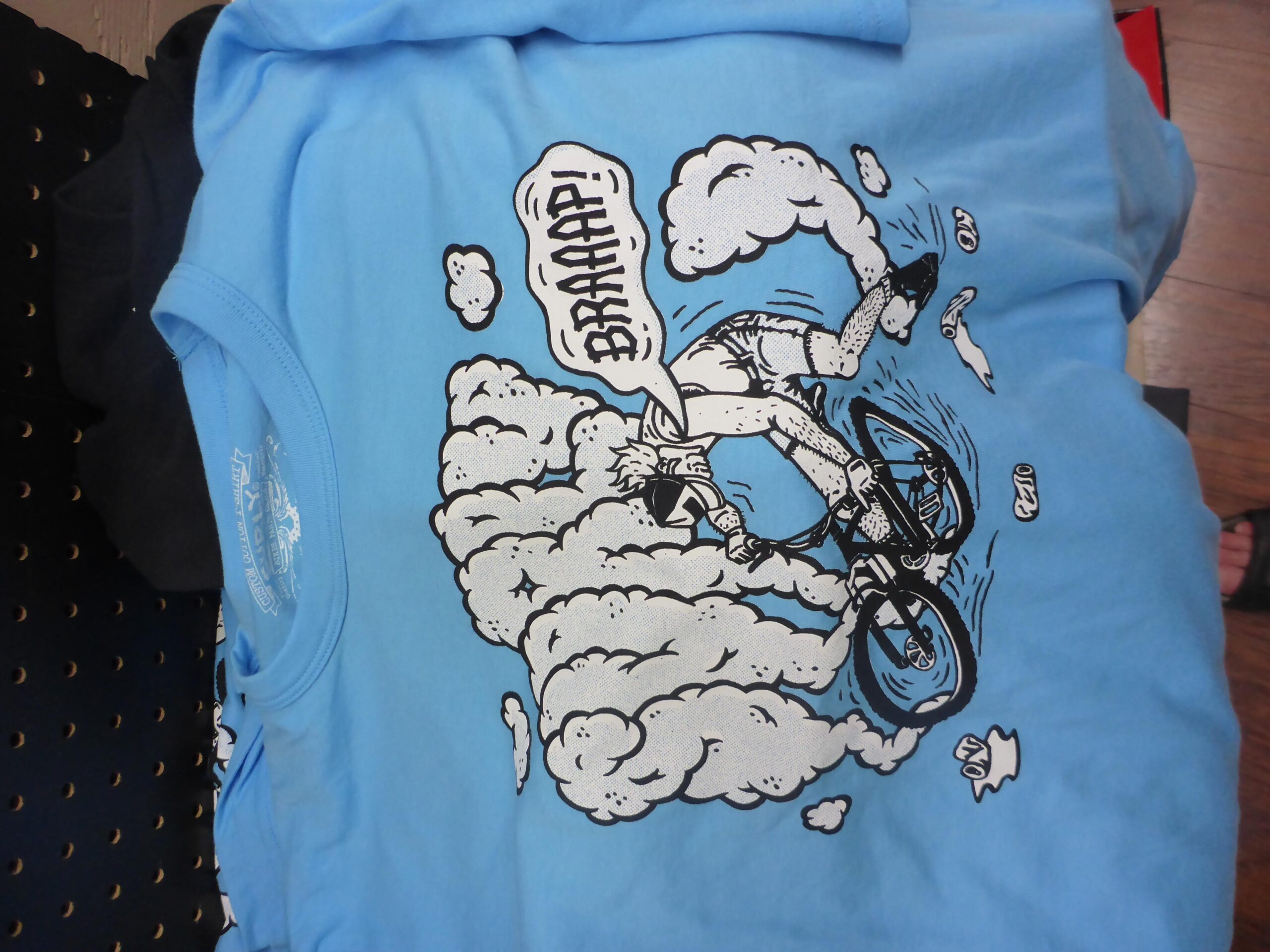 A light blue t-shirt featuring a graphic design of a person riding a bicycle, creating a cloud of dust and smoke. The cyclist is depicted in a dynamic pose, with the text "BRAAAP!" illustrated above. The background includes playful details such as small clouds and scattered items. The shirt is laid flat, partially covering a black fabric underneath.