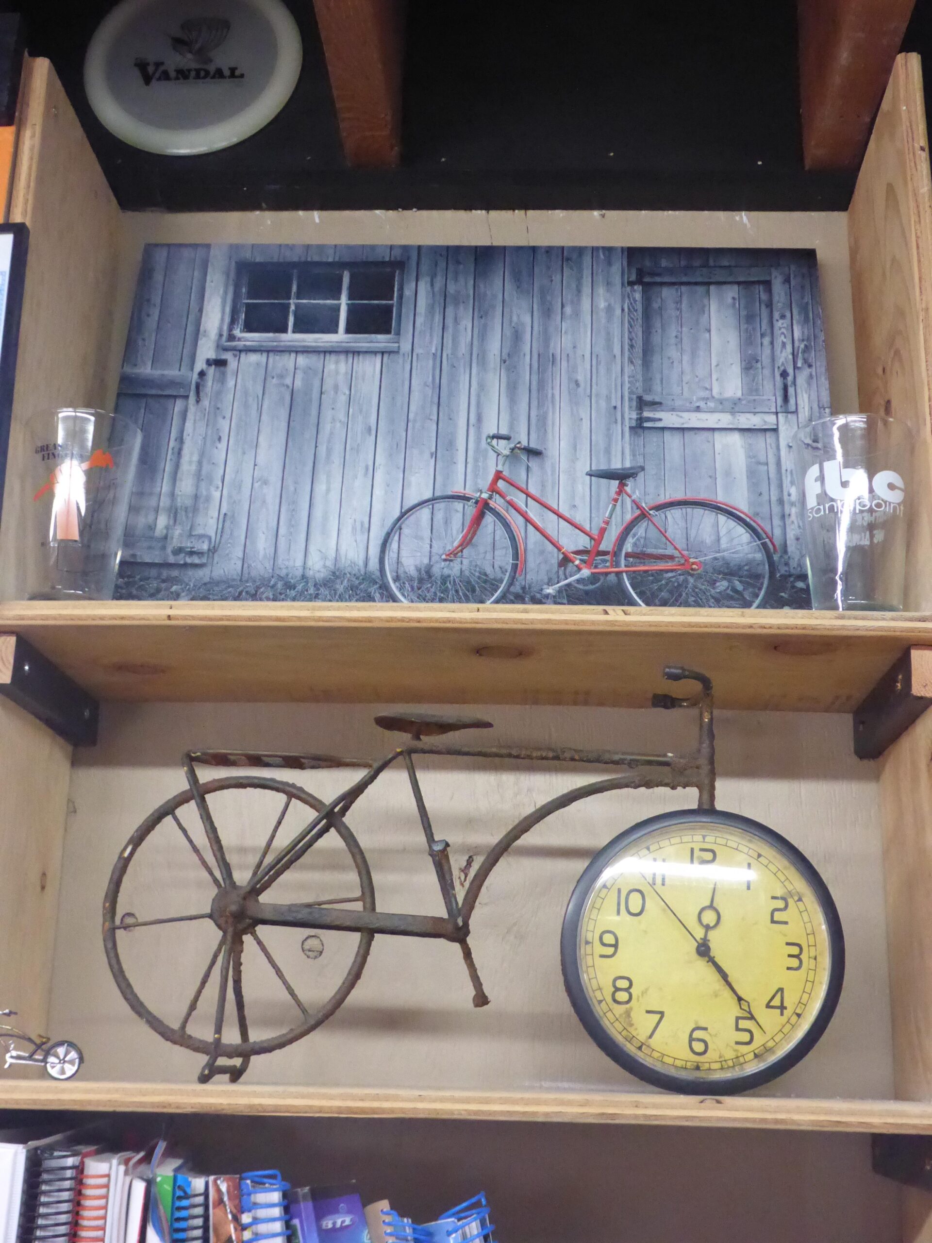 A wooden shelf displaying a vintage theme. On the top shelf, a black-and-white photograph of a rustic building with a red bicycle leaning against it, accompanied by two clear glasses. The lower shelf features a decorative, rusted bicycle sculpture mounted above a round, yellow clock, as well as a small metal toy bicycle. Various notebooks and binders are visible on the bottom shelf.