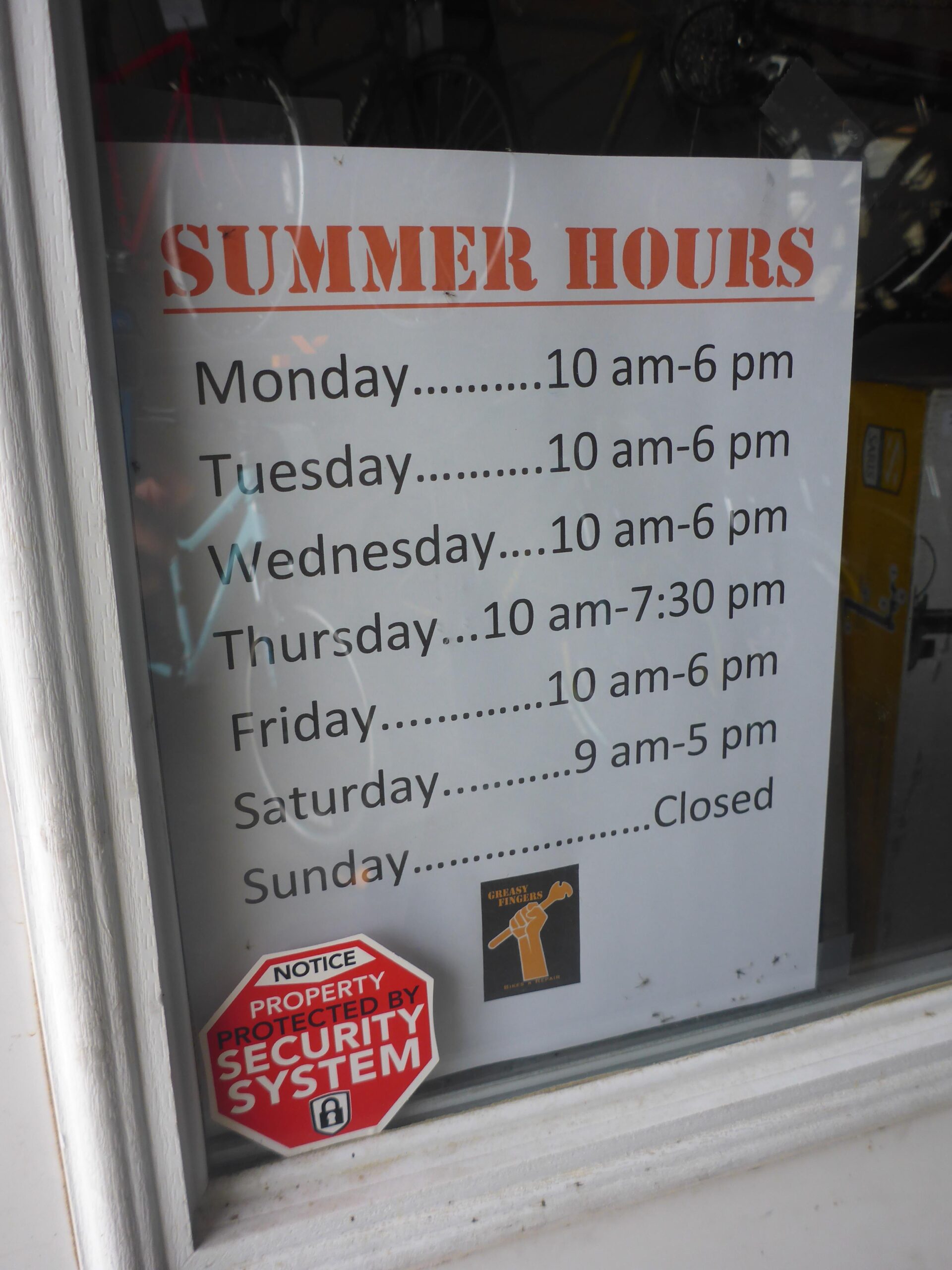 A sign in a window displaying summer hours for a business, with text listing operating times for each day of the week. The sign states the hours as follows: Monday to Wednesday from 10 a.m. to 6 p.m., Thursday from 10 a.m. to 7:30 p.m., Friday from 10 a.m. to 6 p.m., Saturday from 9 a.m. to 5 p.m., and Sunday is closed. A security system notice is visible at the bottom of the sign, along with a logo featuring a wrench and a raised fist.