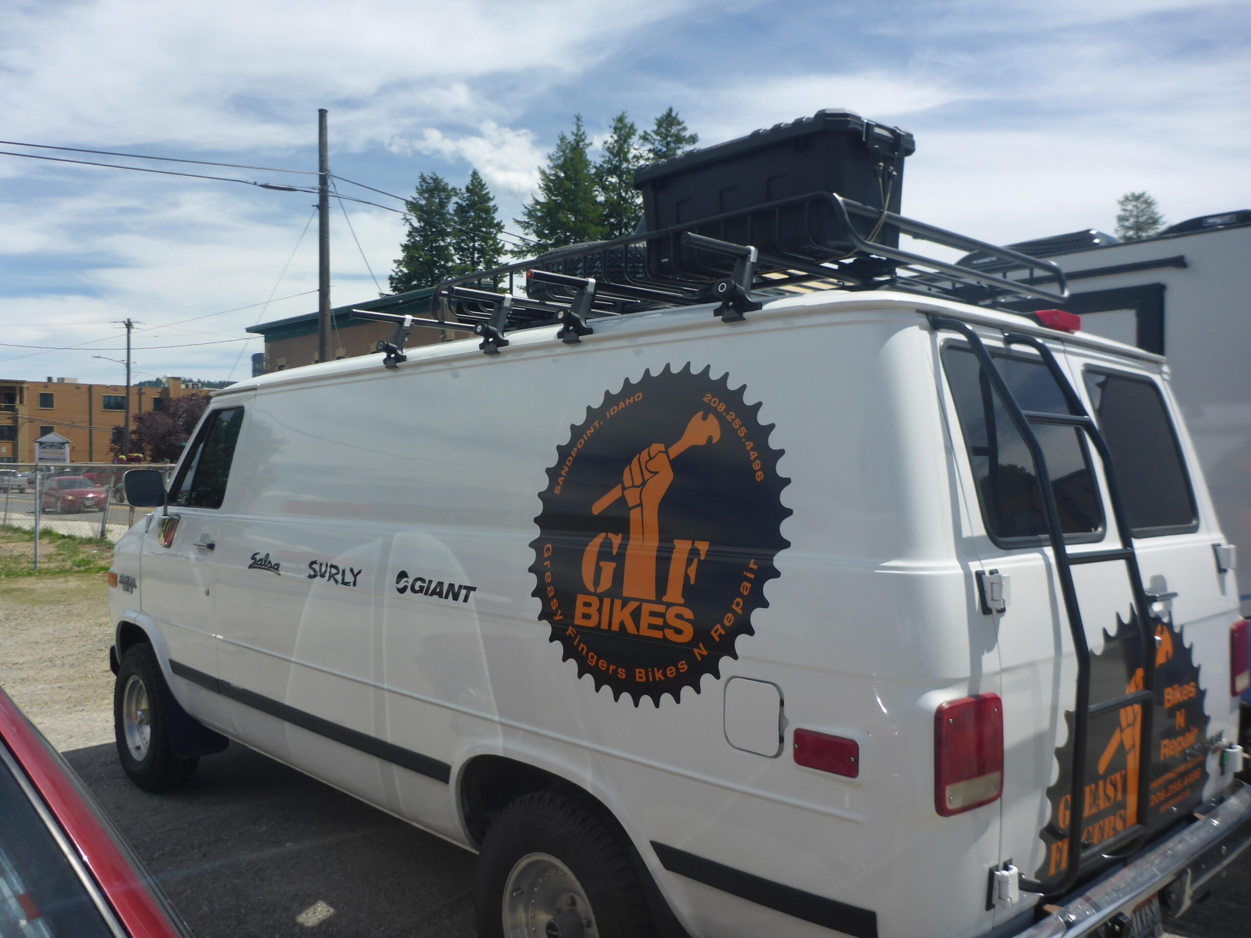 A white van parked in a lot, featuring a large logo on the side that reads "GREAT FINGERS BIKES N REPAIR" along with illustrations of a raised fist and a bicycle gear. The van also displays the names of several bike brands, including Salsa, Surly, and Giant, and has a roof rack with a storage box on top. In the background, there are trees and buildings under a blue sky.