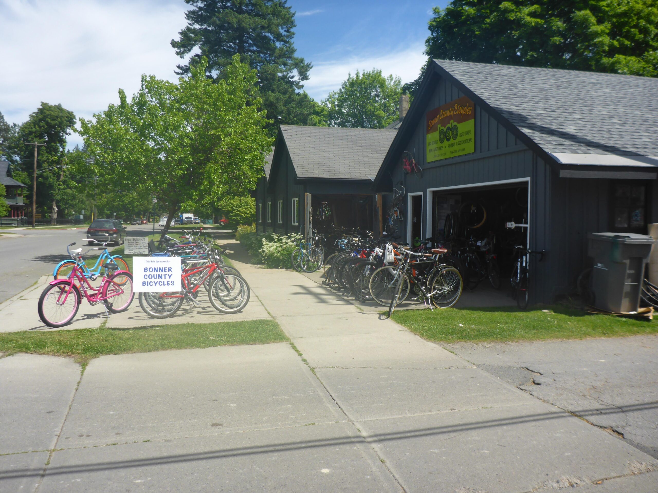 A view of a bicycle shop called "Bonner County Bicycles," featuring a variety of bikes displayed outside. The shop is housed in a dark-colored building with a yellow sign above the entrance. In front, there are two bright bicycles in pink and blue alongside a sign promoting the shop. Trees and a street can be seen in the background, indicating a suburban setting.