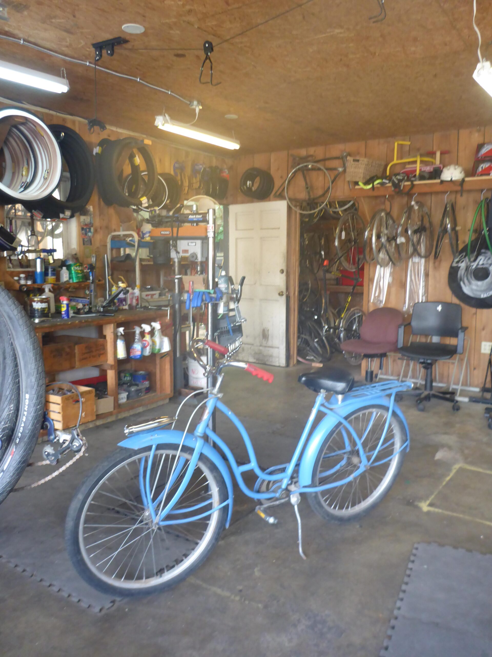 A blue bicycle is parked on the concrete floor of a cluttered garage, surrounded by various bike parts and tools. The walls are lined with hanging bicycle tires and wheels, and a workbench is visible in the background. The garage has overhead lighting and an open door at the back, leading to an outdoor area.