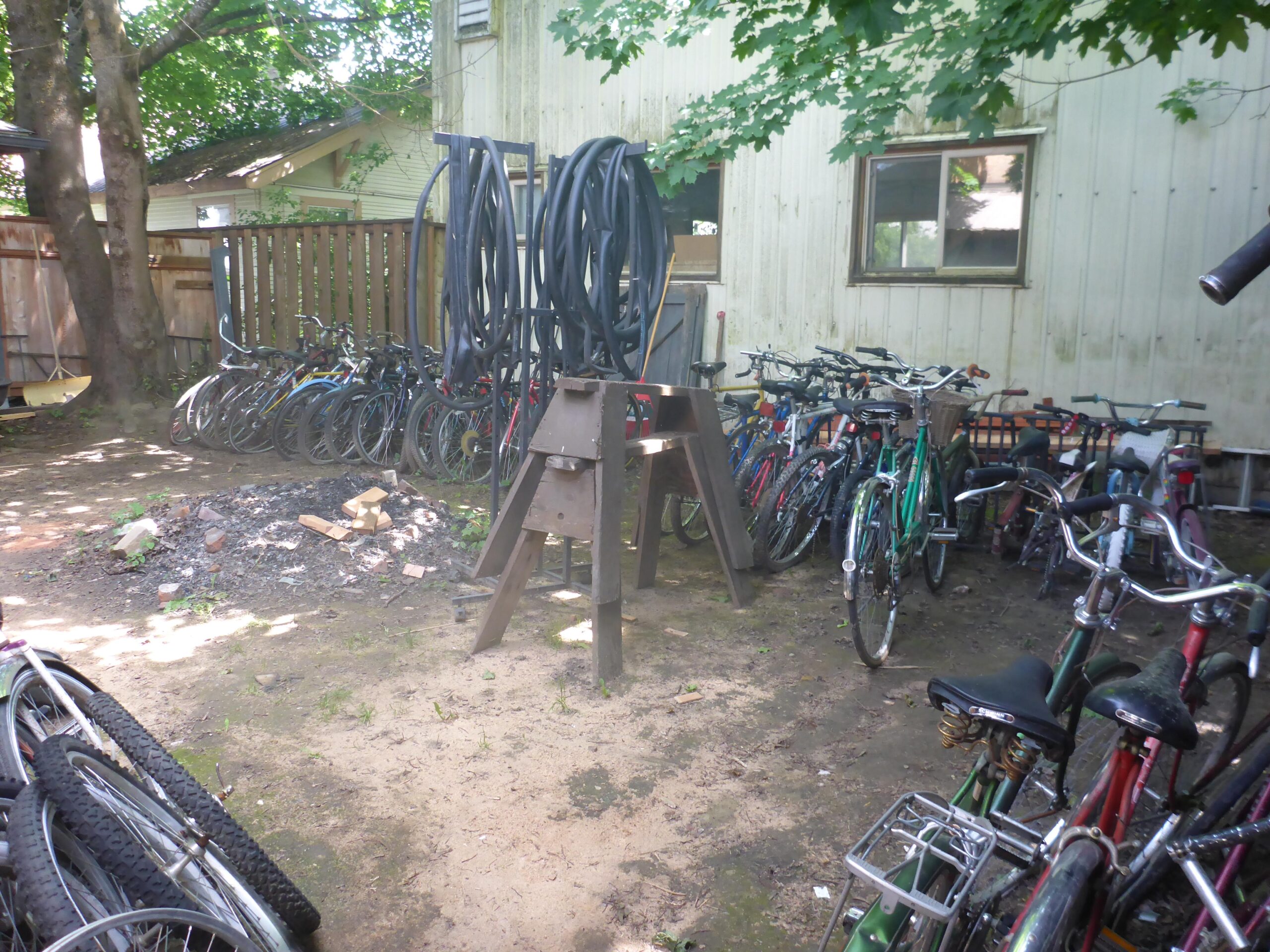 A cluttered backyard scene featuring numerous bicycles of various colors and styles stacked against a building. There are also several large coils of rubber hoses mounted on a frame, and a small area with scattered bricks and debris. The surrounding trees provide dappled sunlight, creating a relaxed outdoor atmosphere.
