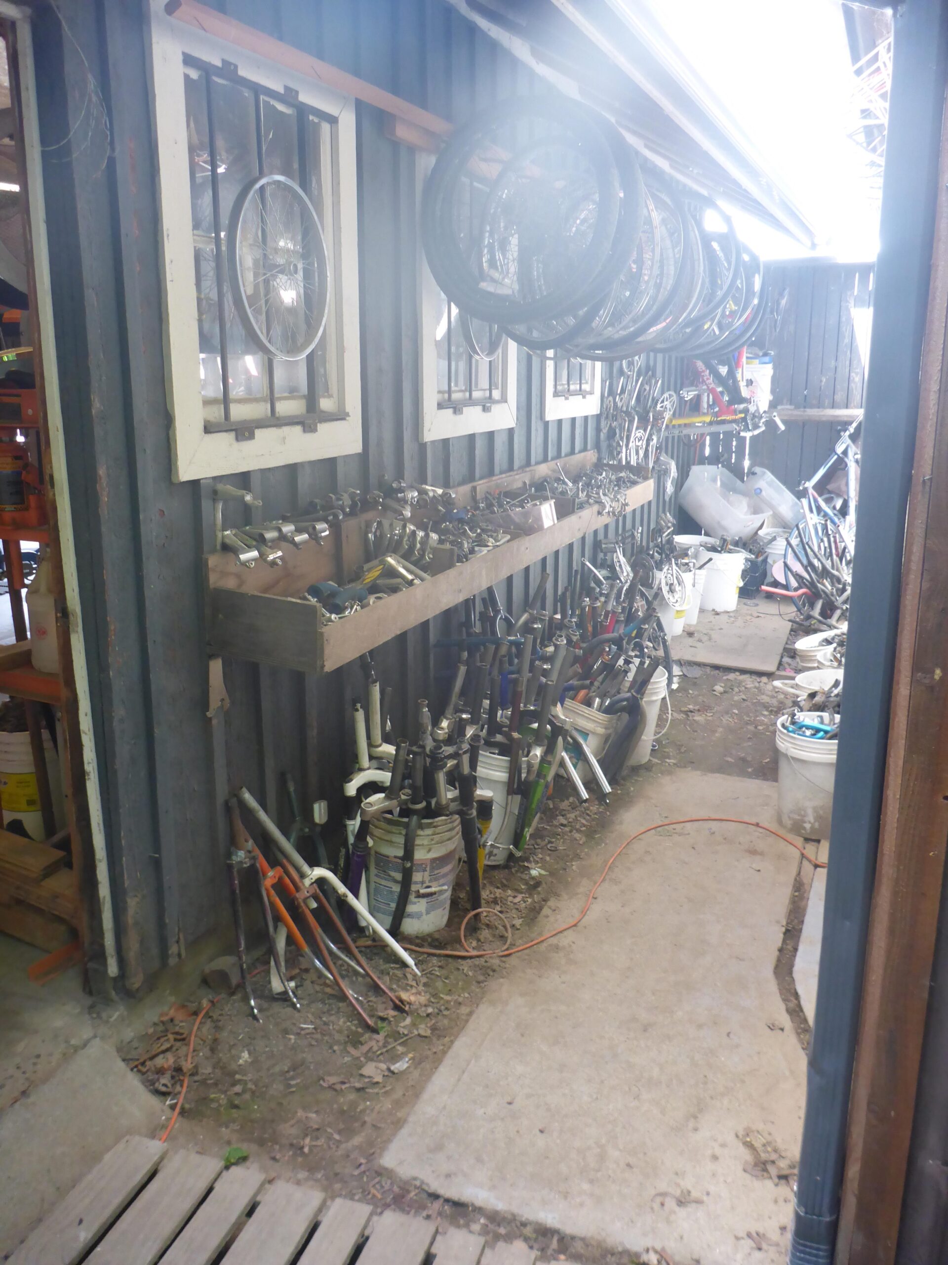 A narrow path leading to a storage area with a dark wooden wall. Several bicycle wheels are hanging from the ceiling, and a wooden shelf holds various bike tools and parts, including forks and handlebars. Buckets filled with bike components are lined up along the side. Sunlight is filtering through the space, creating a bright atmosphere.