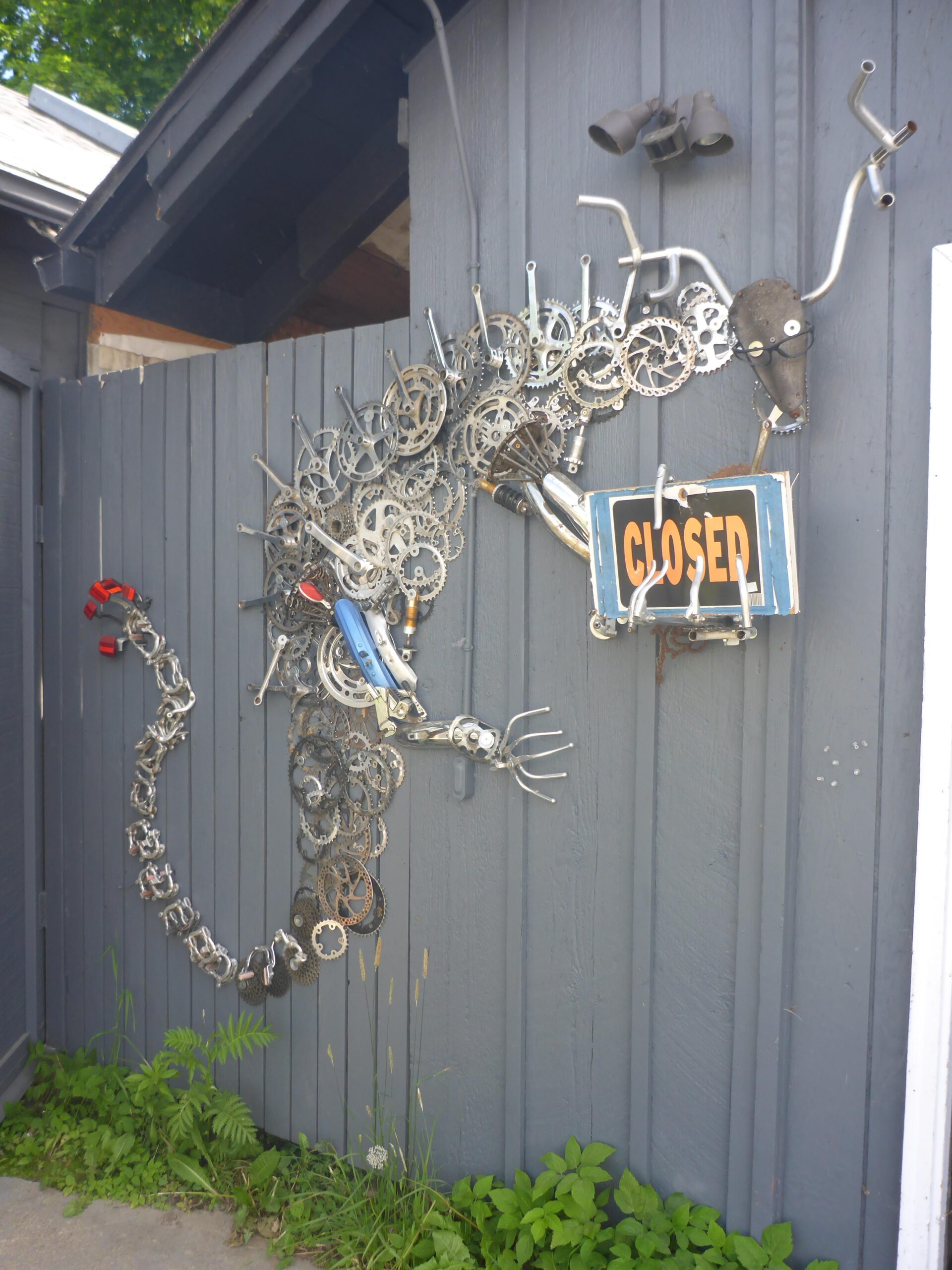 A creative metal sculpture resembling a dragon, made from bicycle parts and other scrap materials, is attached to a grey wooden wall. The sculpture