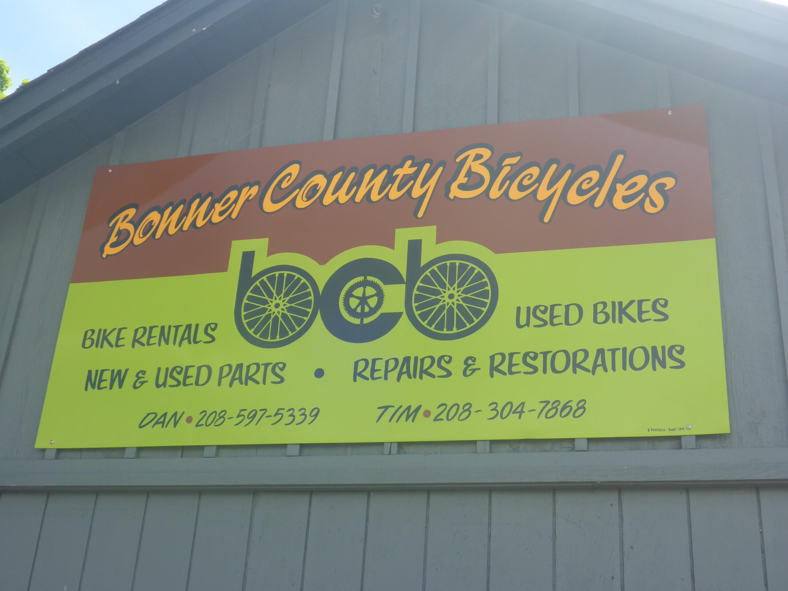 Sign for Bonner County Bicycles featuring services such as bike rentals, new and used parts, and bike repairs and restorations. Contact information is provided for two individuals. The sign has a colorful design with a mix of green and brown backgrounds.