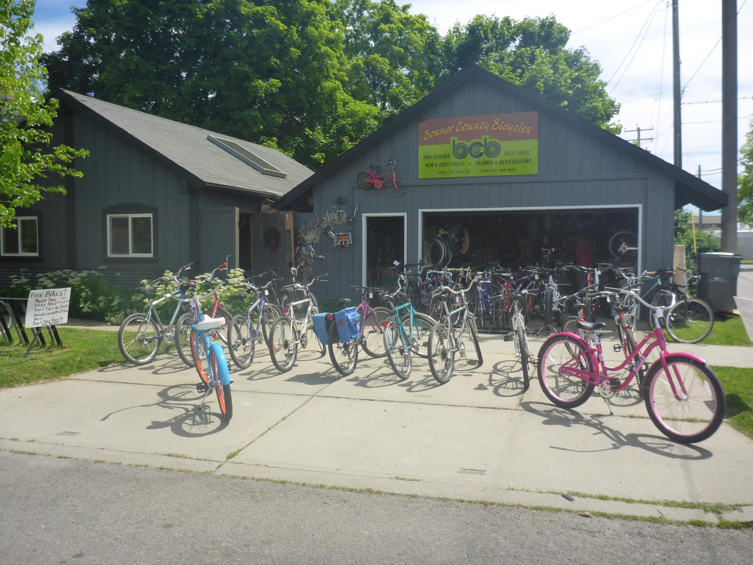 A quaint bike shop with a gray wooden exterior, featuring a large sign that reads "Bonner County Bicycles." In front, several bicycles of various colors and styles are parked on the concrete driveway. A sign beside the entrance indicates that free bikes are available. The background includes green trees and power lines under a clear blue sky.