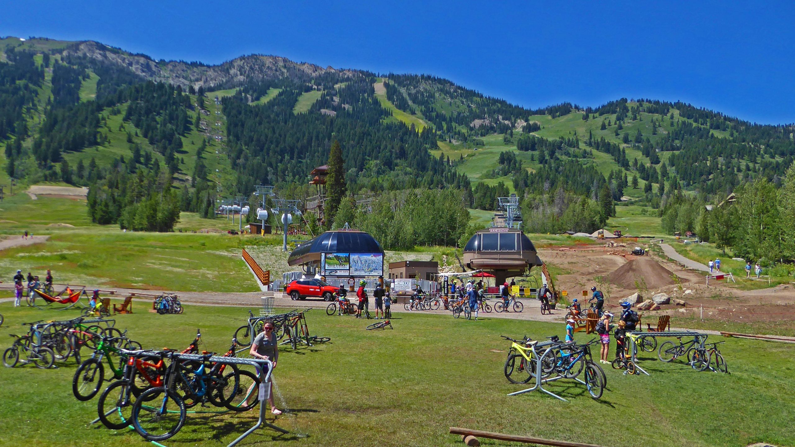 A scenic outdoor area featuring a grassy field filled with bicycles parked on racks, surrounded by trees and mountains in the background. People are enjoying various activities, including biking and relaxing in hammocks. In the center, there are buildings with signs, ski lifts in operation, and a clear blue sky overhead. Jackson Hole Bike Park mountain bike trail.
