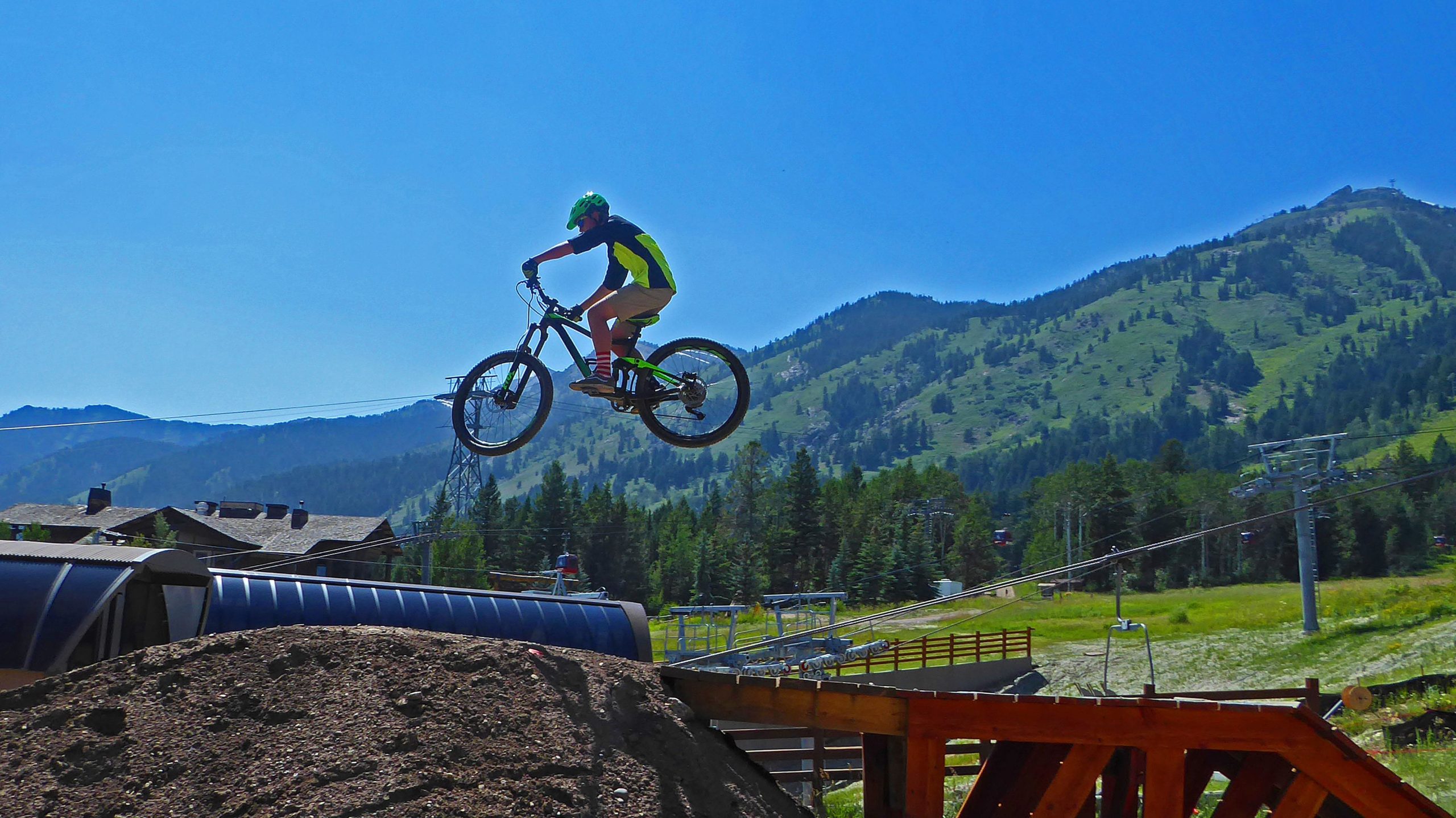 A young cyclist in a green helmet and bright clothing performs a jump on a mountain bike over a rocky ramp, set against a backdrop of lush green mountains and a clear blue sky. Jackson Hole Bike Park mountain bike trail.