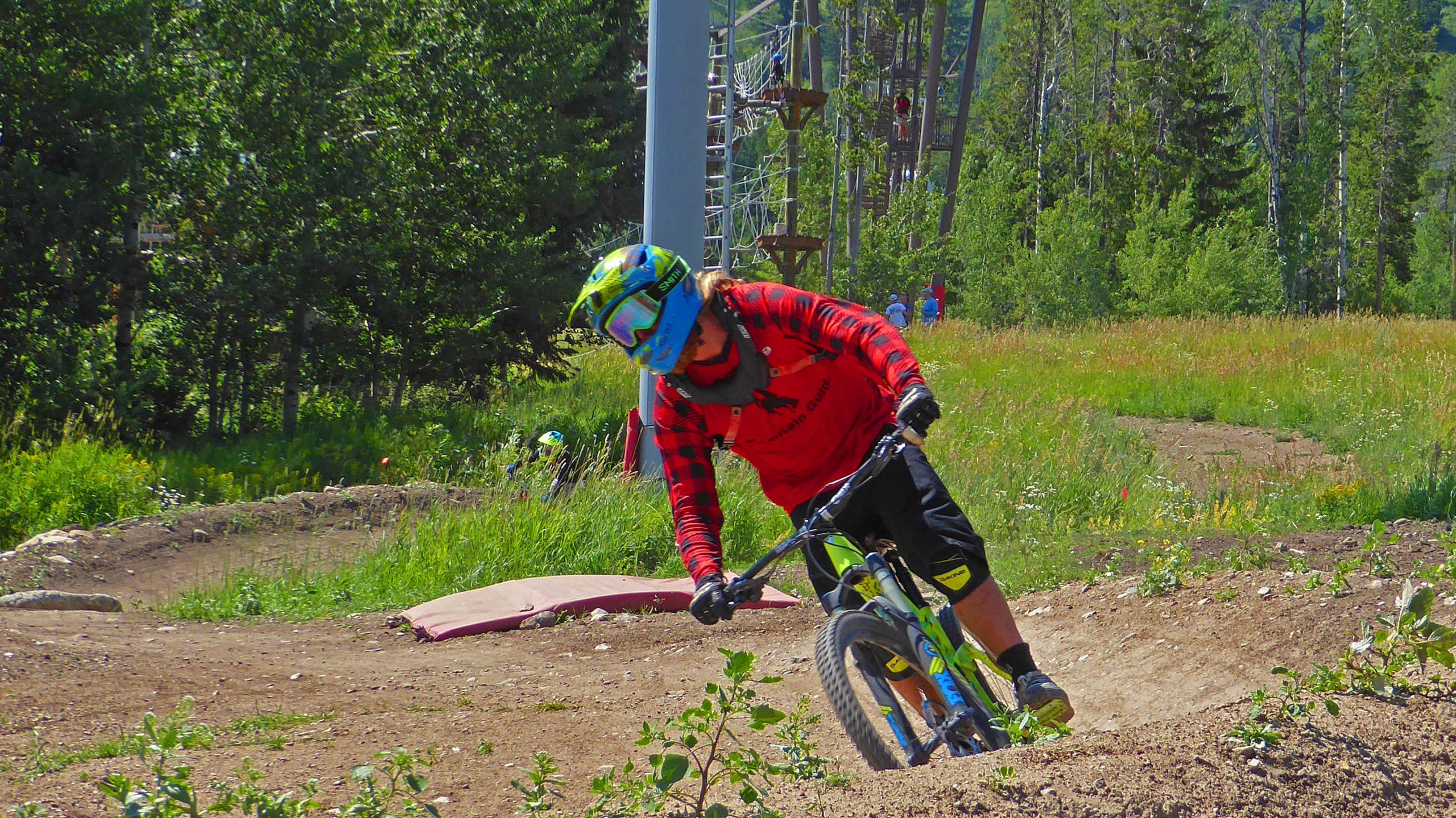 A mountain biker in a red shirt and helmet rides on a dirt trail through a grassy area, leaning into a turn. Trees and a ski lift are visible in the background, along with other bikers in the distance. Jackson Hole Bike Park mountain bike trail.