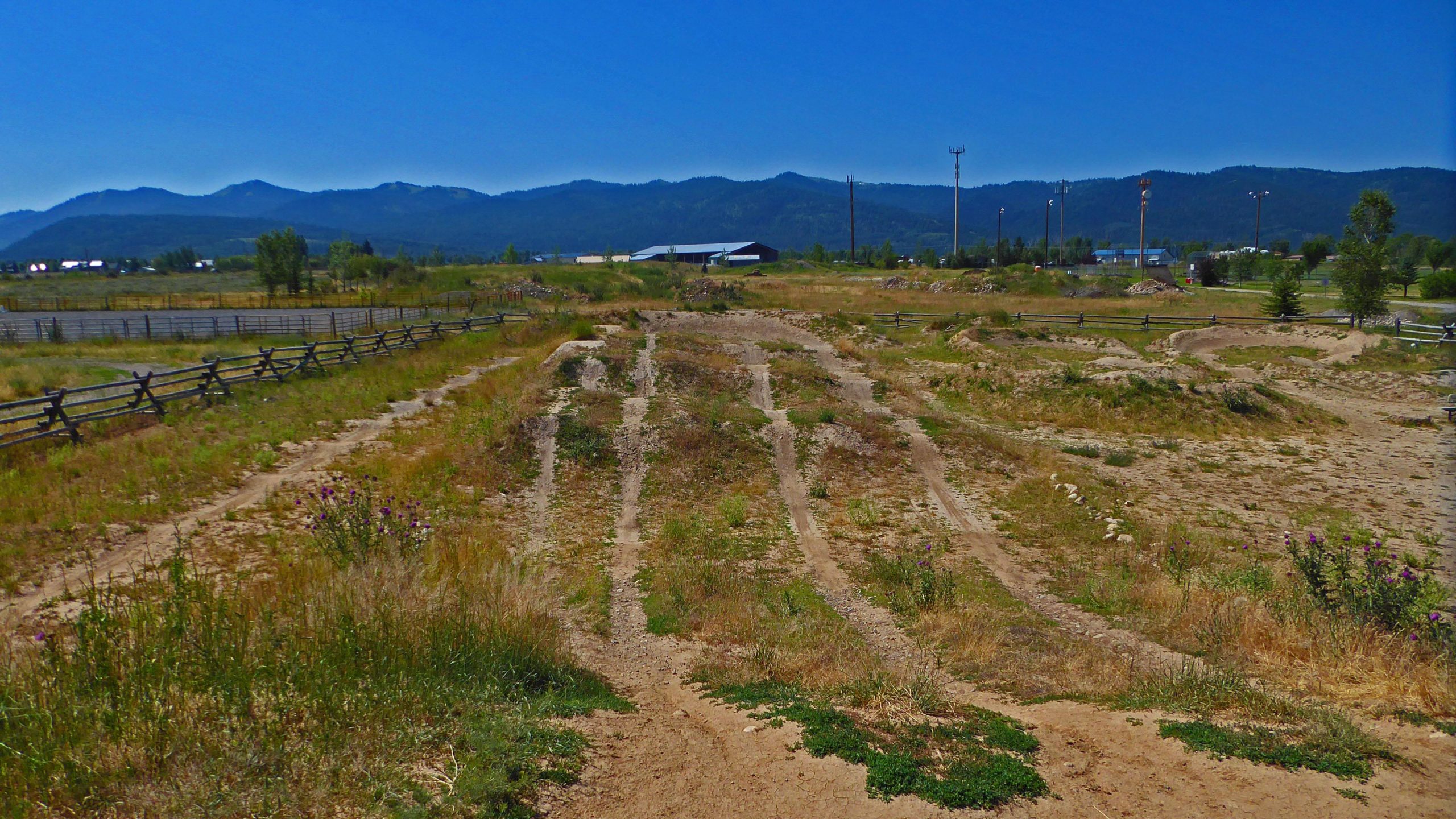 A wide view of a dirt bike track with tire marks, surrounded by tall grass and wildflowers. In the background, there are rolling hills and mountains under a clear blue sky. Various structures and trees are visible along the horizon, with a wooden fence bordering part of the area. Victor Bike Park mountain bike trail.