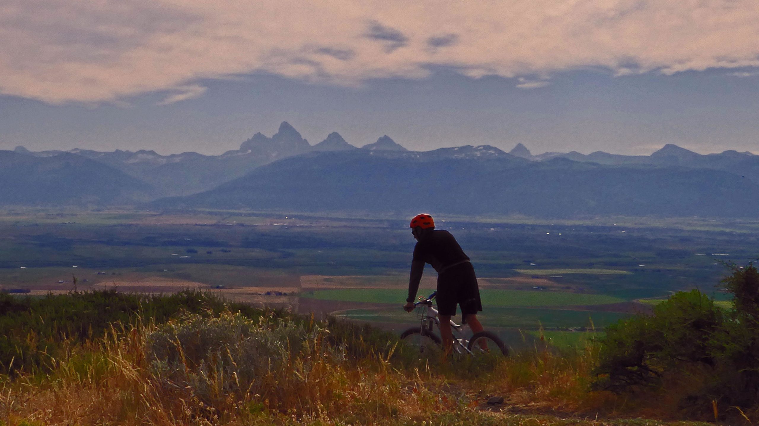 A cyclist in a black outfit and orange helmet stands beside a mountain bike, gazing out over a scenic view of mountains and valleys. The landscape features lush green fields and distant jagged peaks under a cloudy sky. Southbound mountain bike trail.