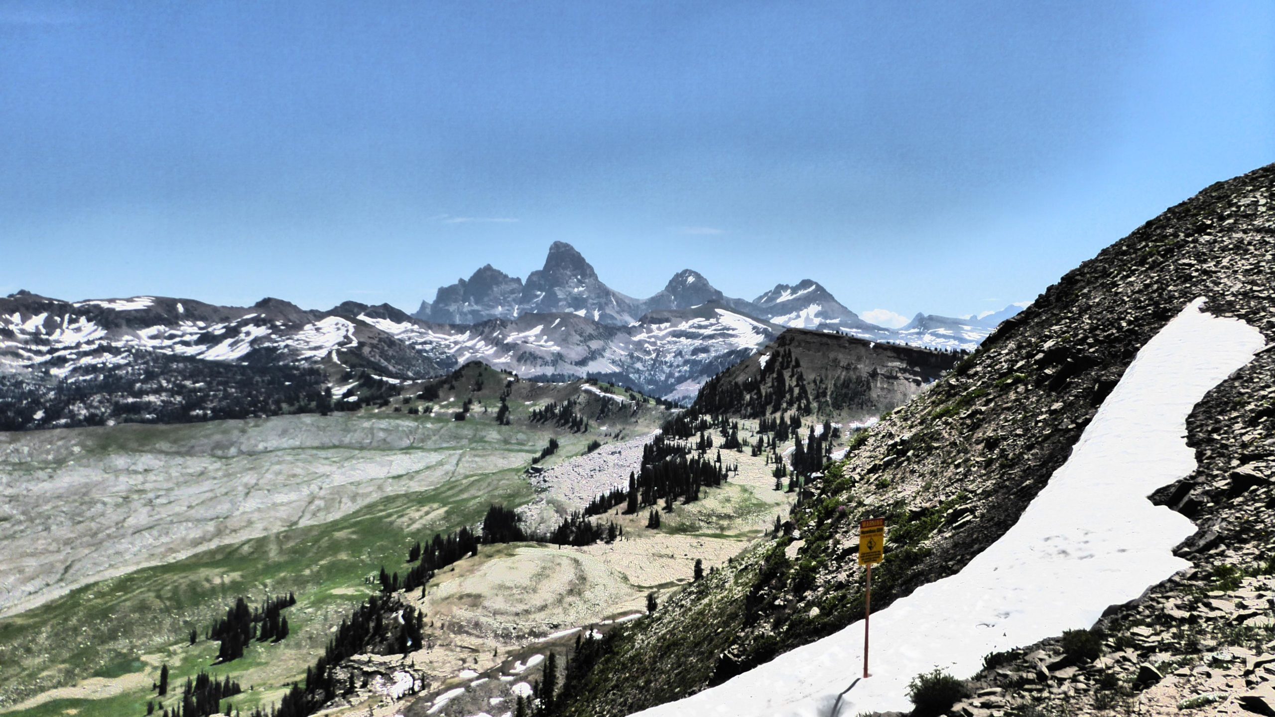A panoramic view of a mountainous landscape featuring rugged peaks in the distance, some capped with snow. The foreground includes rocky terrain with patches of snow and green vegetation. A warning sign is visible along the trail, indicating potential hazards. The sky is clear and blue, enhancing the bright, natural scenery. Grand Targhee Bike Park mountain bike trail.