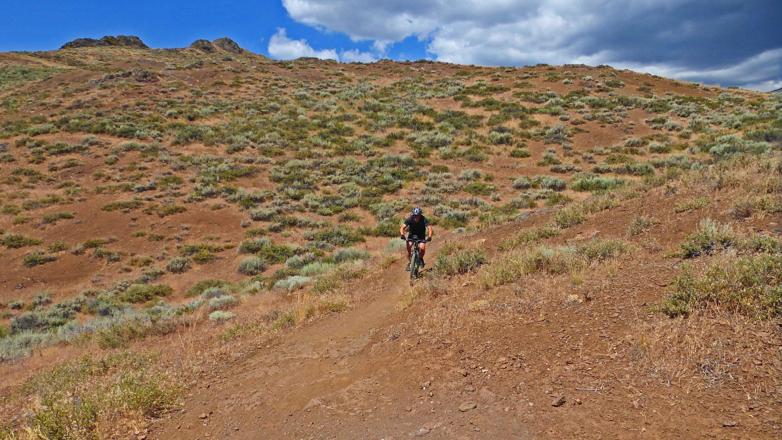 A mountain biker navigating a dirt trail on a hillside covered in sparse vegetation and rocky terrain, under a partly cloudy blue sky. Bullion Connector mountain bike trail.