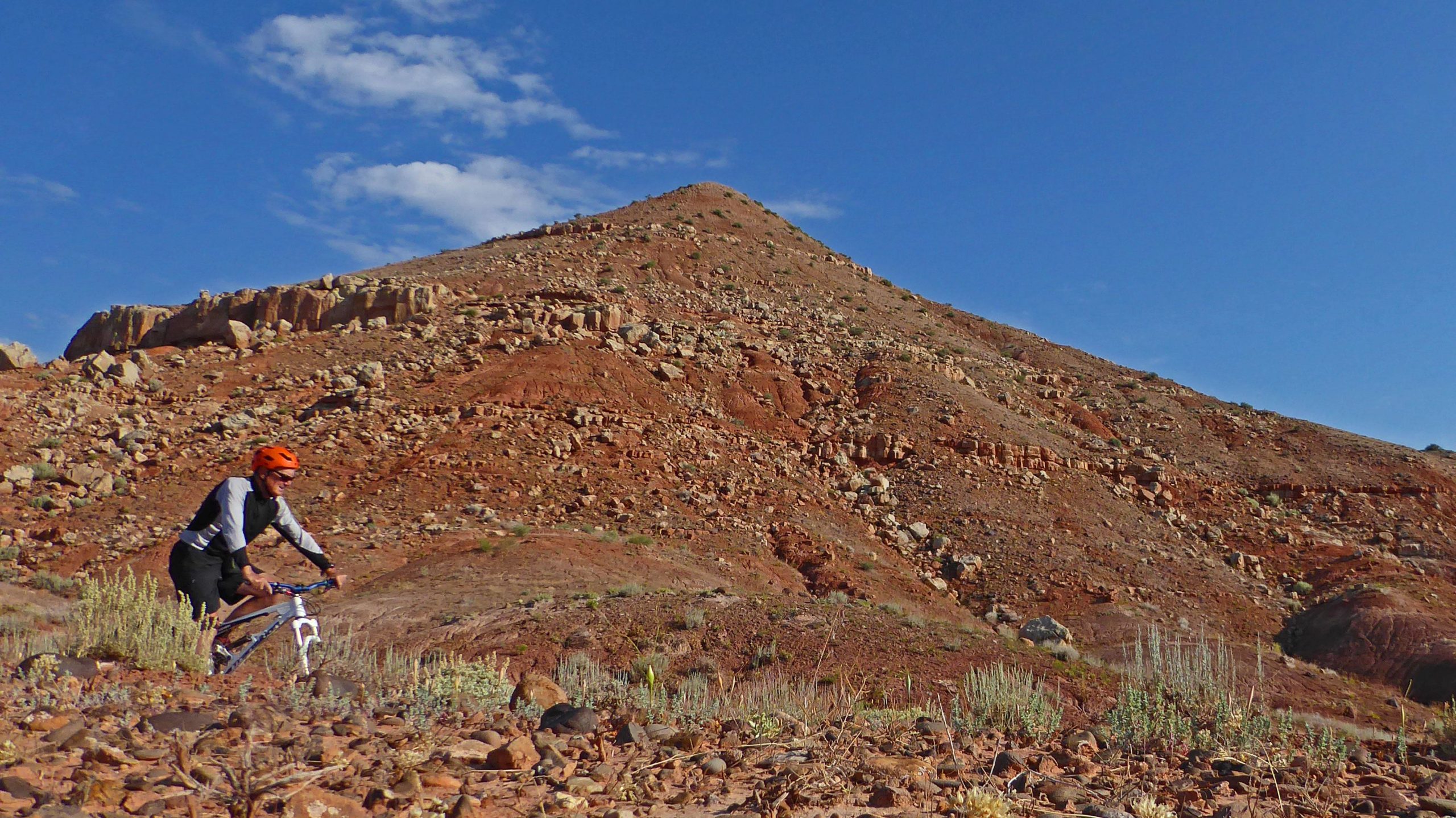 A mountain biker riding along a rocky trail in a arid landscape, with a steep red hillside and a blue sky in the background. McCoy Flat Trails mountain bike trail.