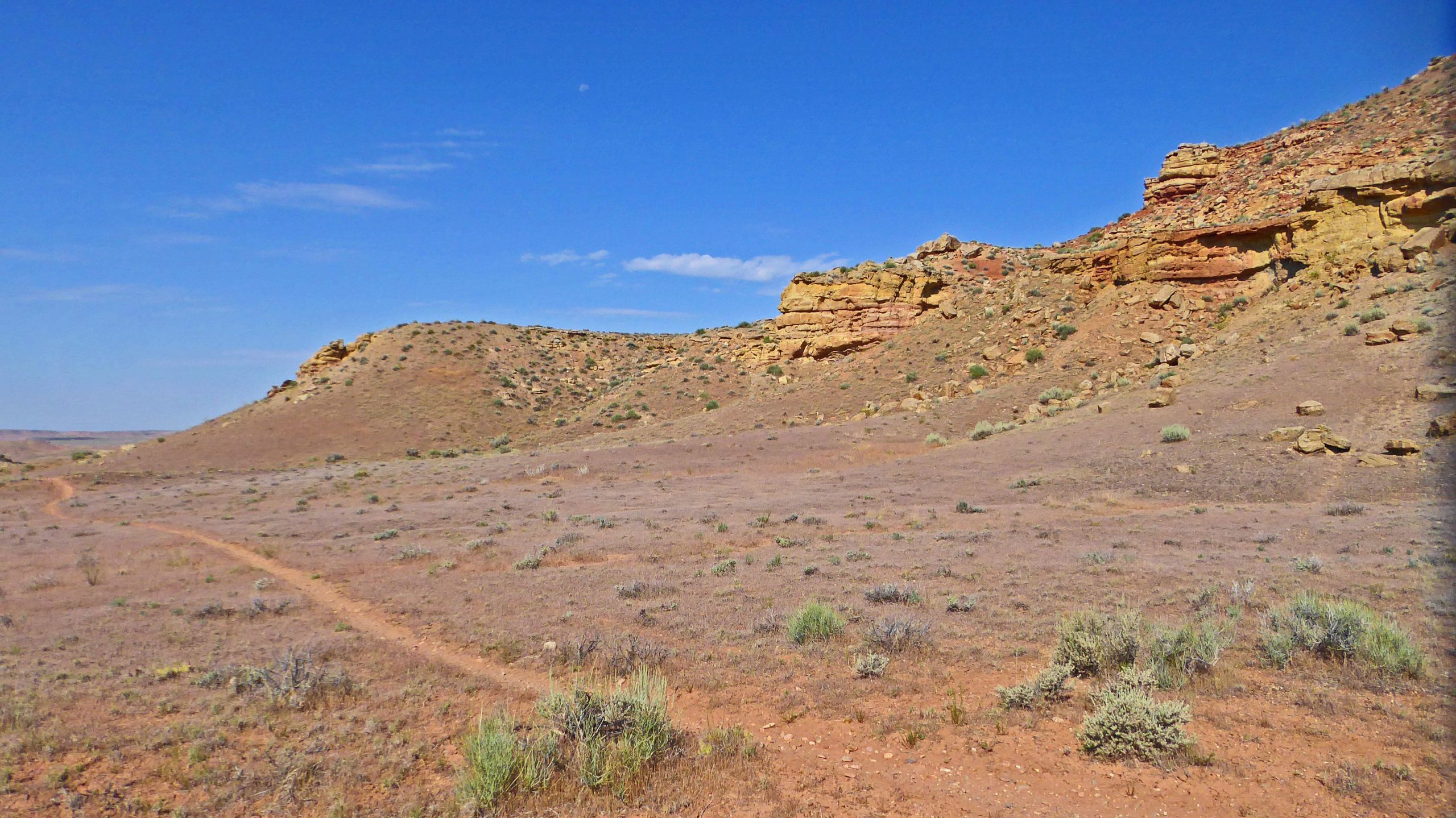A scenic view of a dry, rocky landscape featuring rolling hills with exposed rock formations and sparse vegetation under a clear blue sky. A winding dirt path leads through the foreground, inviting exploration of the arid terrain. McCoy Flat Trails mountain bike trail.