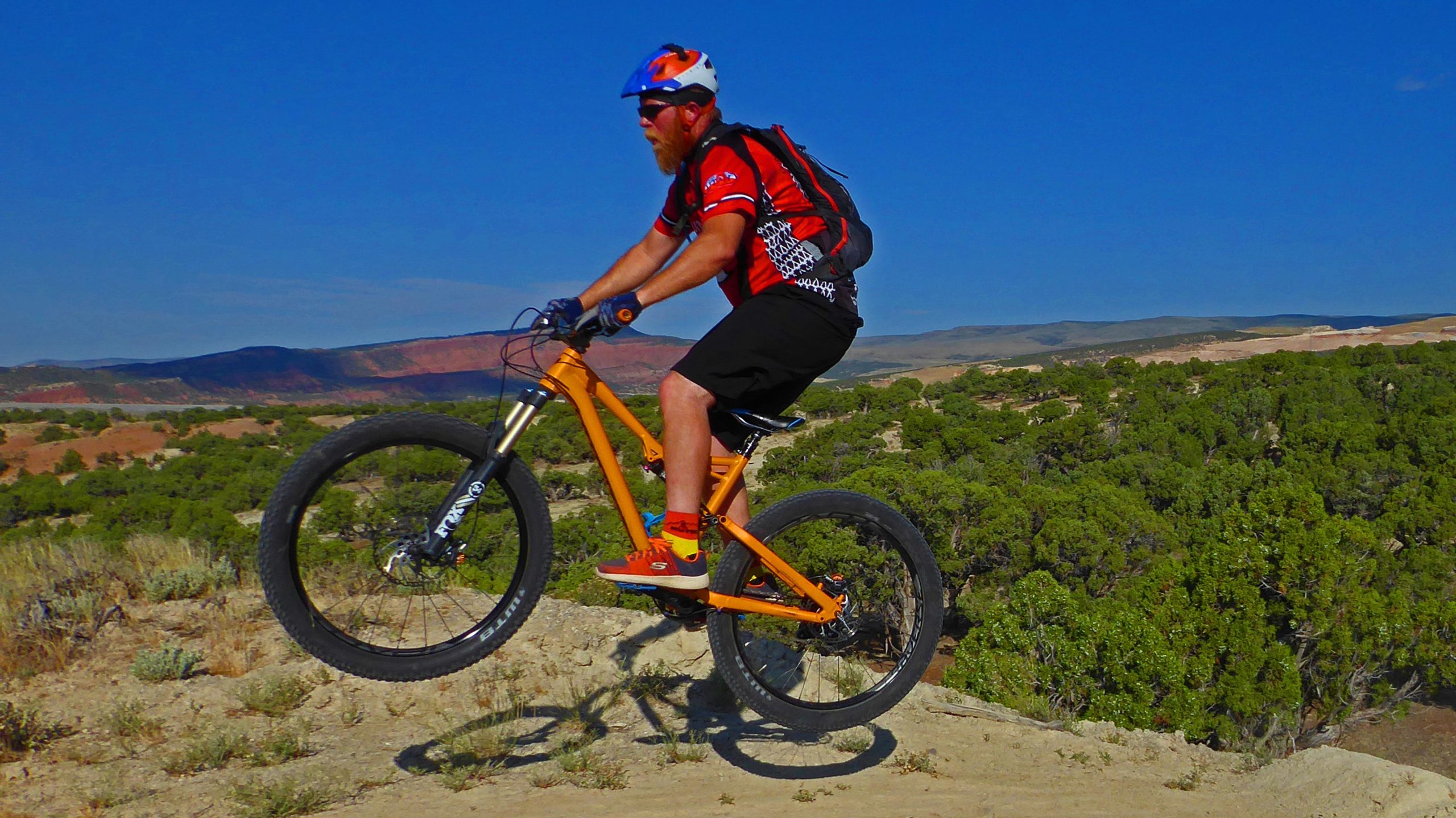 A cyclist performing a jump on a mountain bike in a rugged outdoor landscape, with a backdrop of hills and greenery under a clear blue sky. The rider is wearing a helmet, sunglasses, and a colorful jersey, showcasing an active and adventurous spirit. Jazz Chrome Molly mountain bike trail.