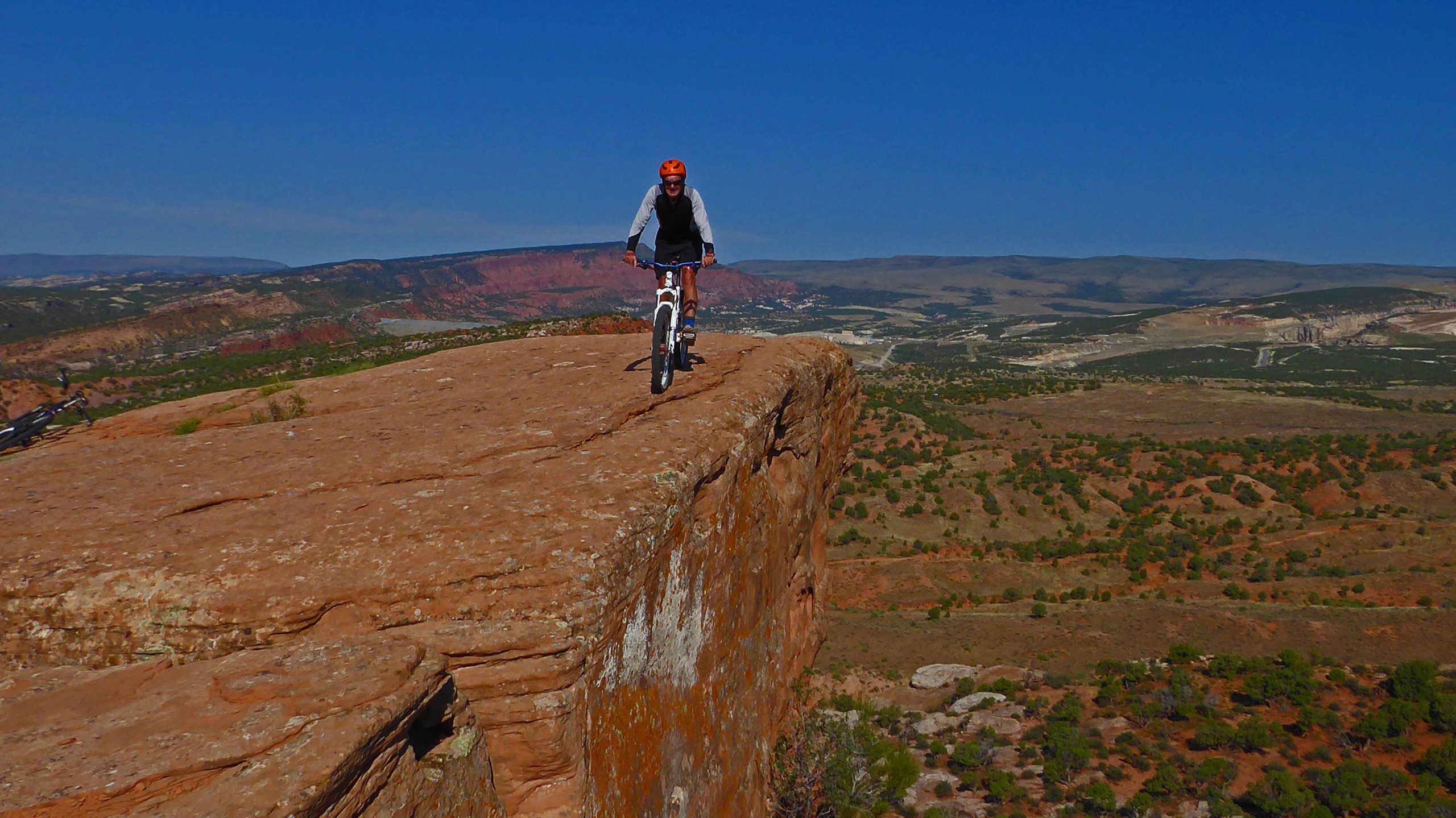 A mountain biker navigating along the edge of a rocky cliff, with a panoramic view of a vast landscape featuring hills, roads, and shrubs under a clear blue sky. J-Boy mountain bike trail.