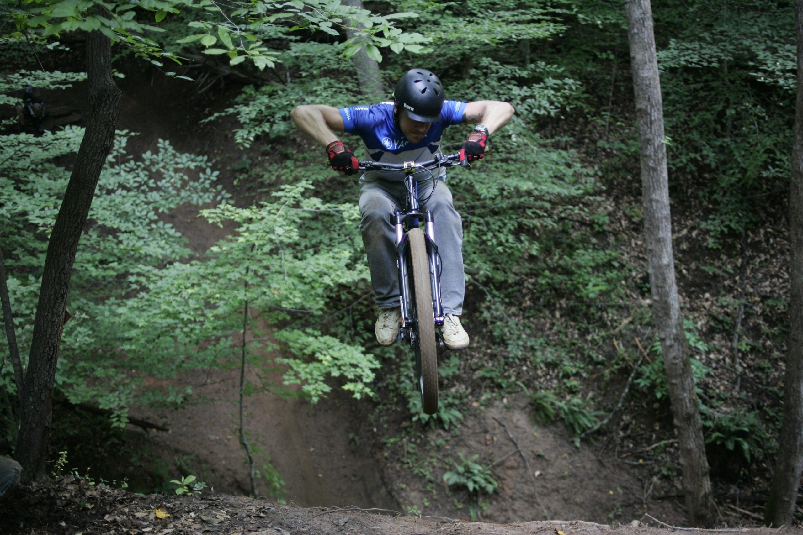 A mountain biker performing a jump off a dirt ramp in a forested area, surrounded by lush green trees. The rider, wearing a helmet, blue shirt, and gloves, is suspended in mid-air above the ground, showcasing dynamic movement and skill. Salem Lake mountain bike trail.