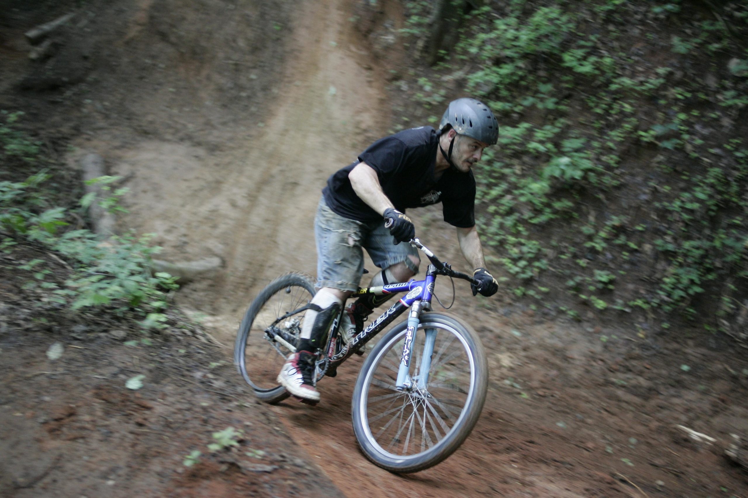 A mountain biker navigating a dirt trail, leaning into a turn while wearing a helmet, gloves, and shorts, surrounded by greenery and a sloped, earthy terrain. Salem Lake mountain bike trail.