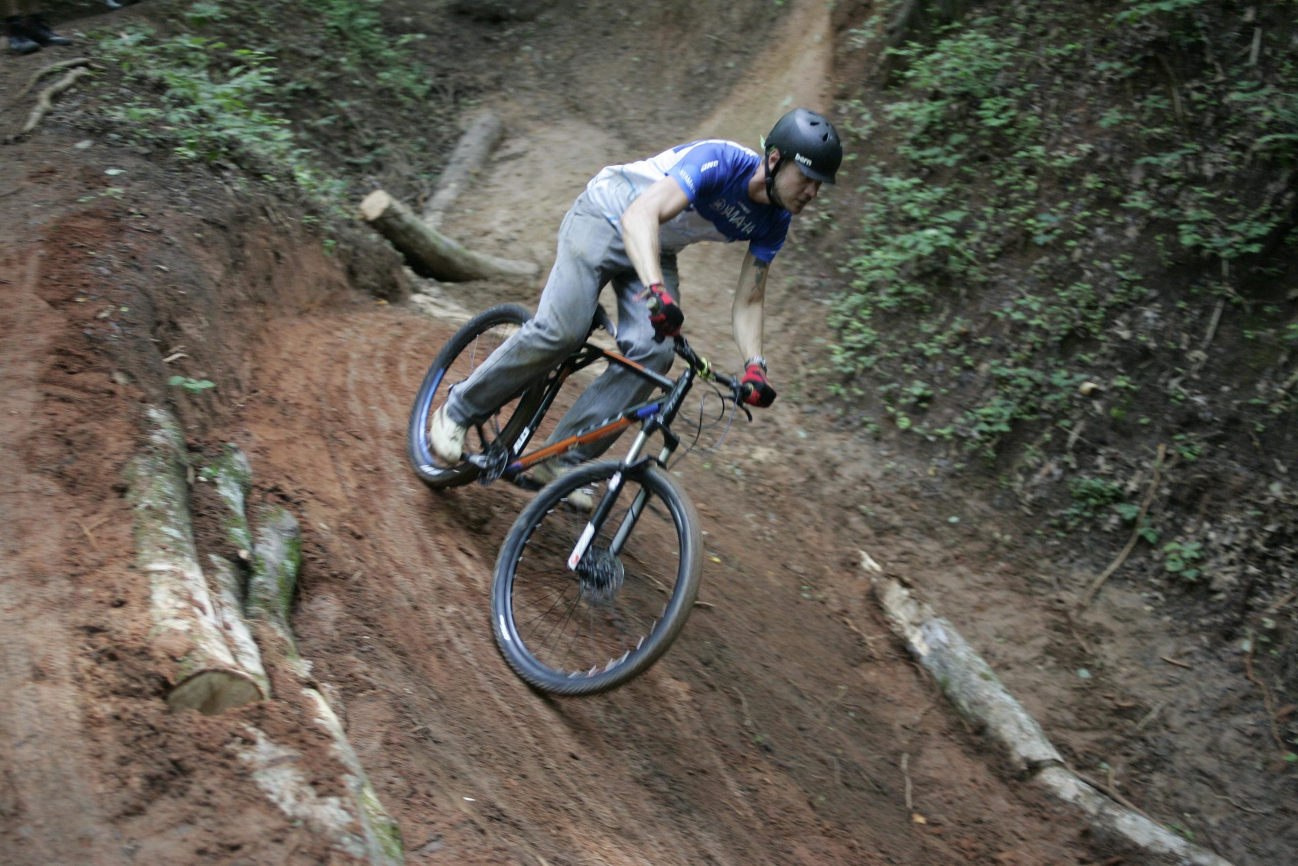 A mountain biker riding down a dirt trail, navigating through a curve with trees and logs on either side. The rider is wearing a helmet, a blue shirt, and gloves, and appears to be focused on maintaining balance on the bike. Salem Lake mountain bike trail.