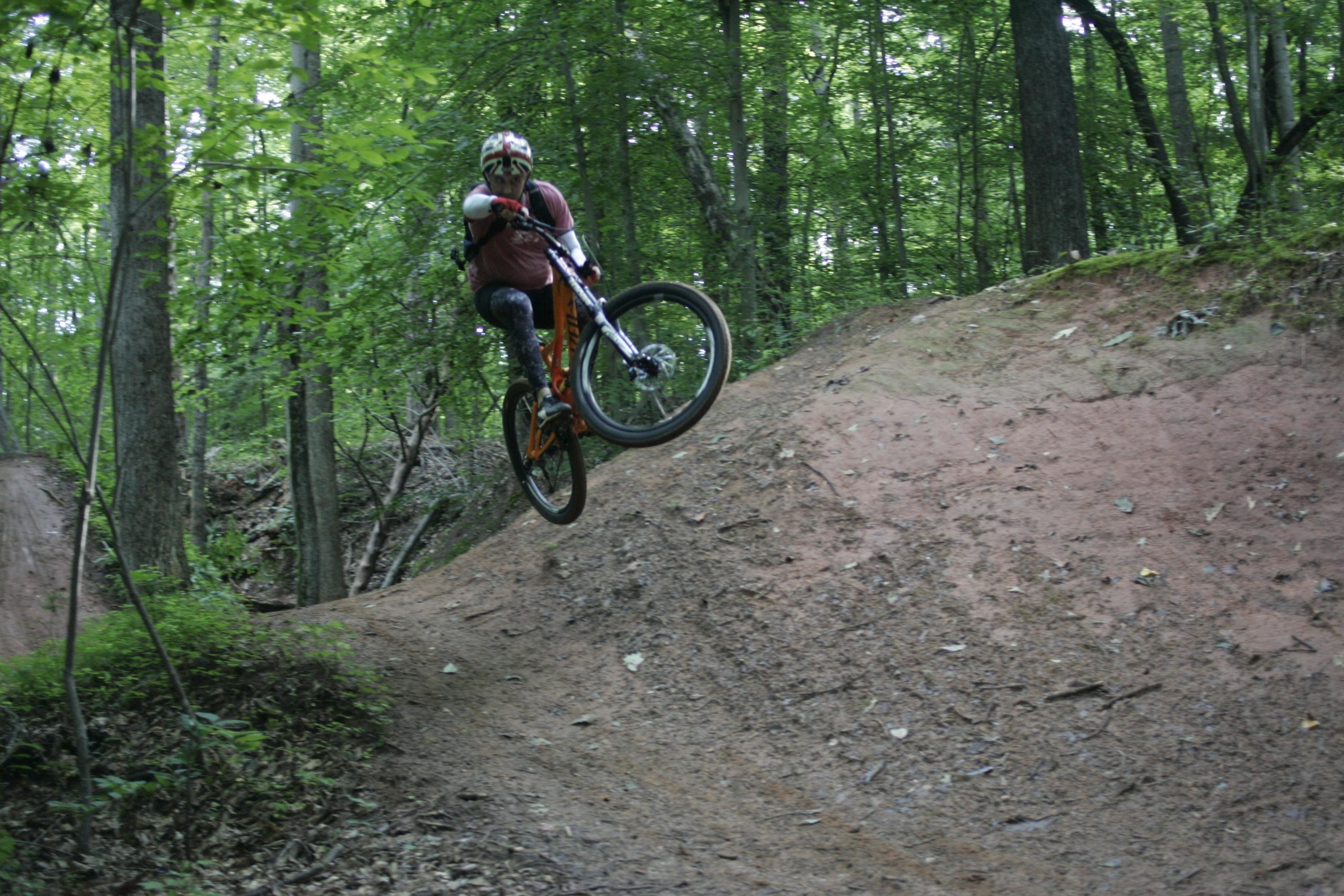 A mountain biker performs a jump off a dirt ramp in a forested area, surrounded by lush green trees. The biker is wearing protective gear and an orange bicycle, with the bike's front wheel elevated above the ground while airborne. The scene captures a dynamic moment of action and outdoor sports. Salem Lake mountain bike trail.