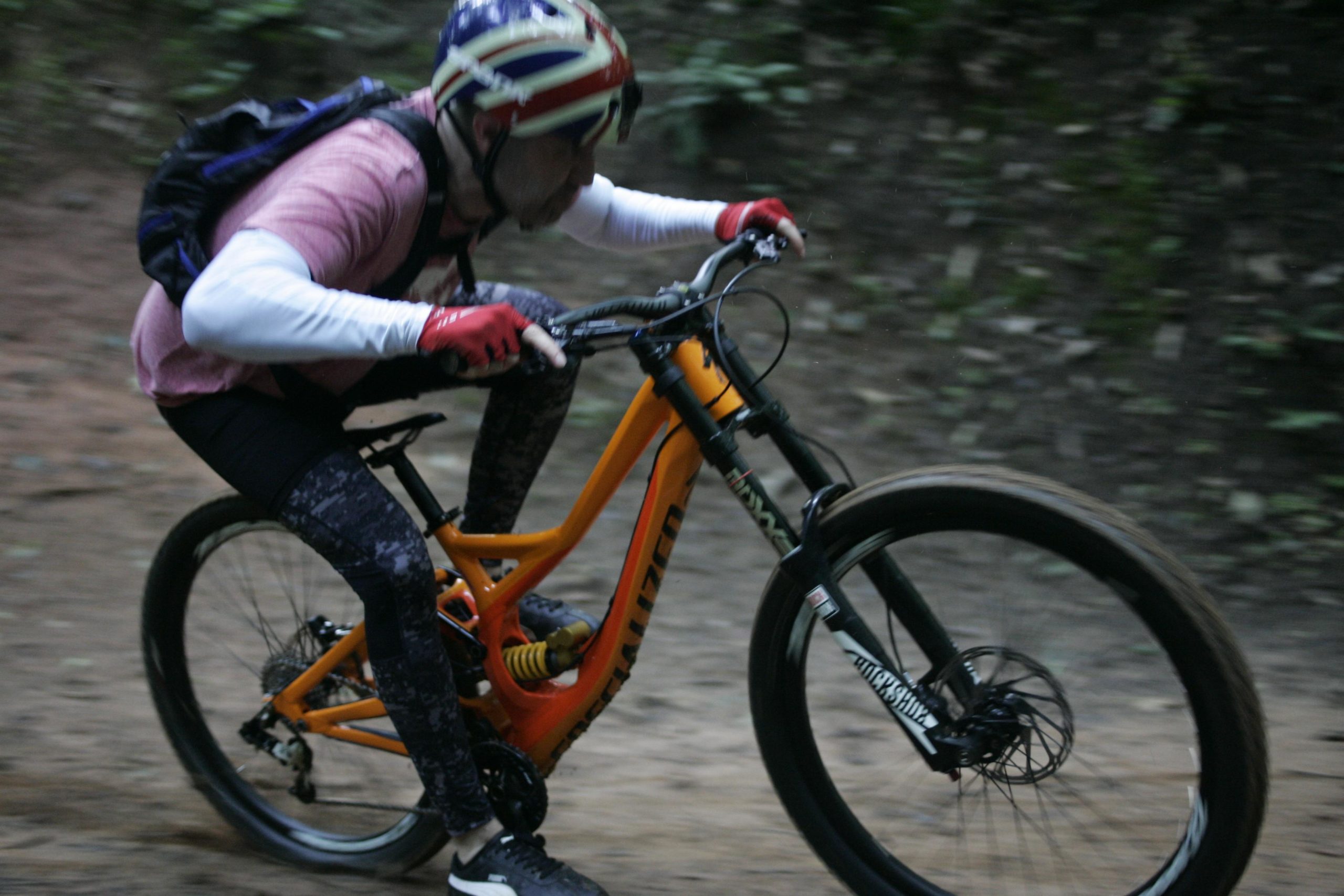 A person wearing a helmet and gloves rides a mountain bike down a forested trail. They are crouched low, with focus on navigating the terrain, as the orange bike contrasts against the earthy background. Salem Lake mountain bike trail.