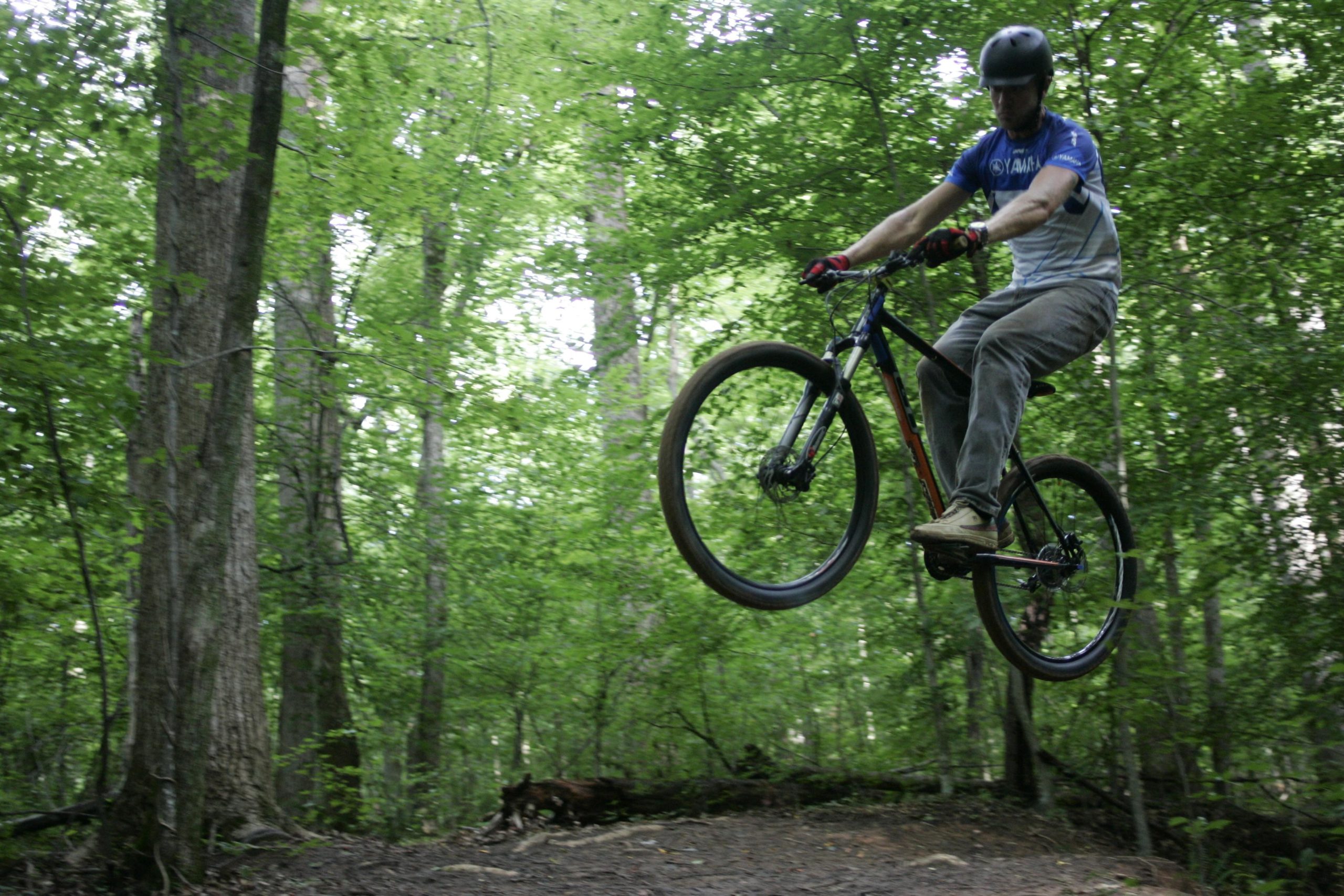 A mountain biker performing a jump on a dirt trail surrounded by lush green trees in a forest. The biker is wearing a helmet and cycling gloves, showcasing an action-packed moment of adventure in nature. Salem Lake mountain bike trail.