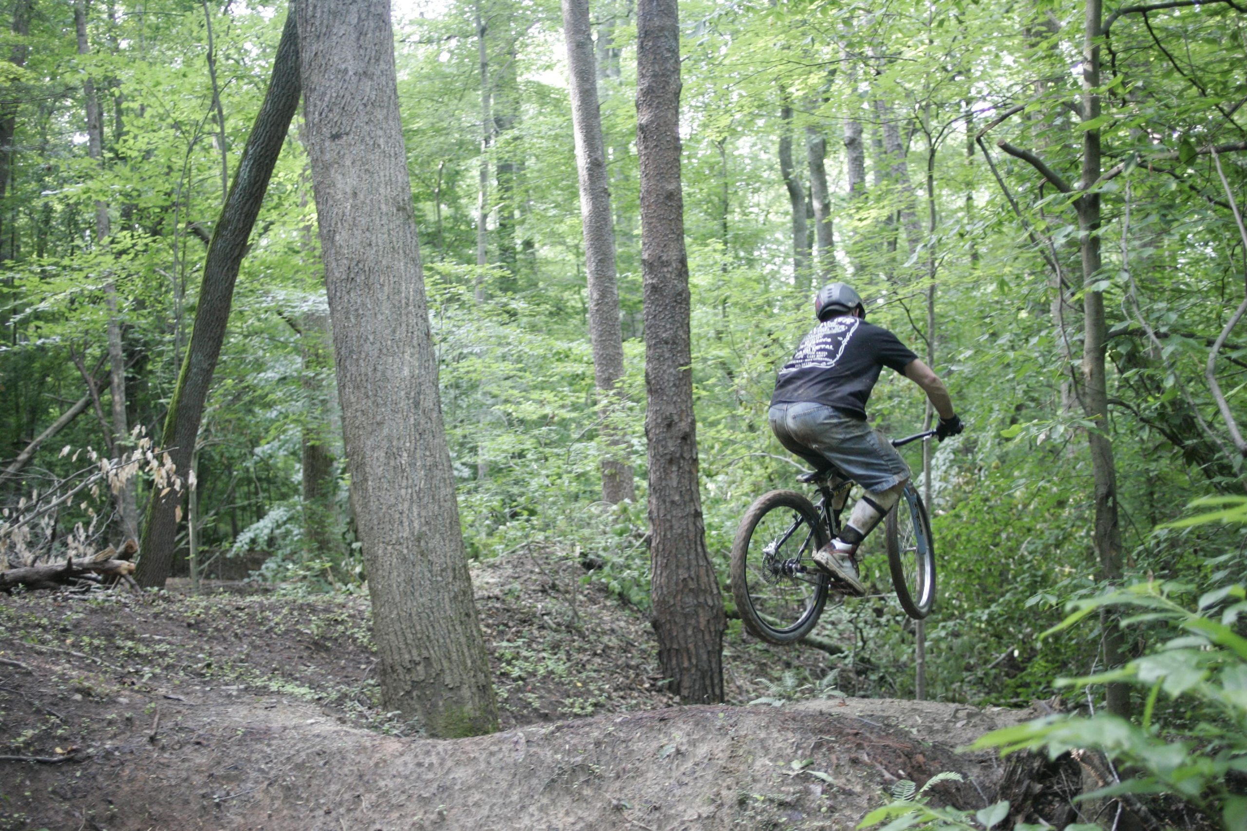 A mountain biker performing a jump on a dirt trail surrounded by dense green trees and foliage. The rider is wearing a helmet and casual cycling attire, with sunlight filtering through the trees. Salem Lake mountain bike trail.