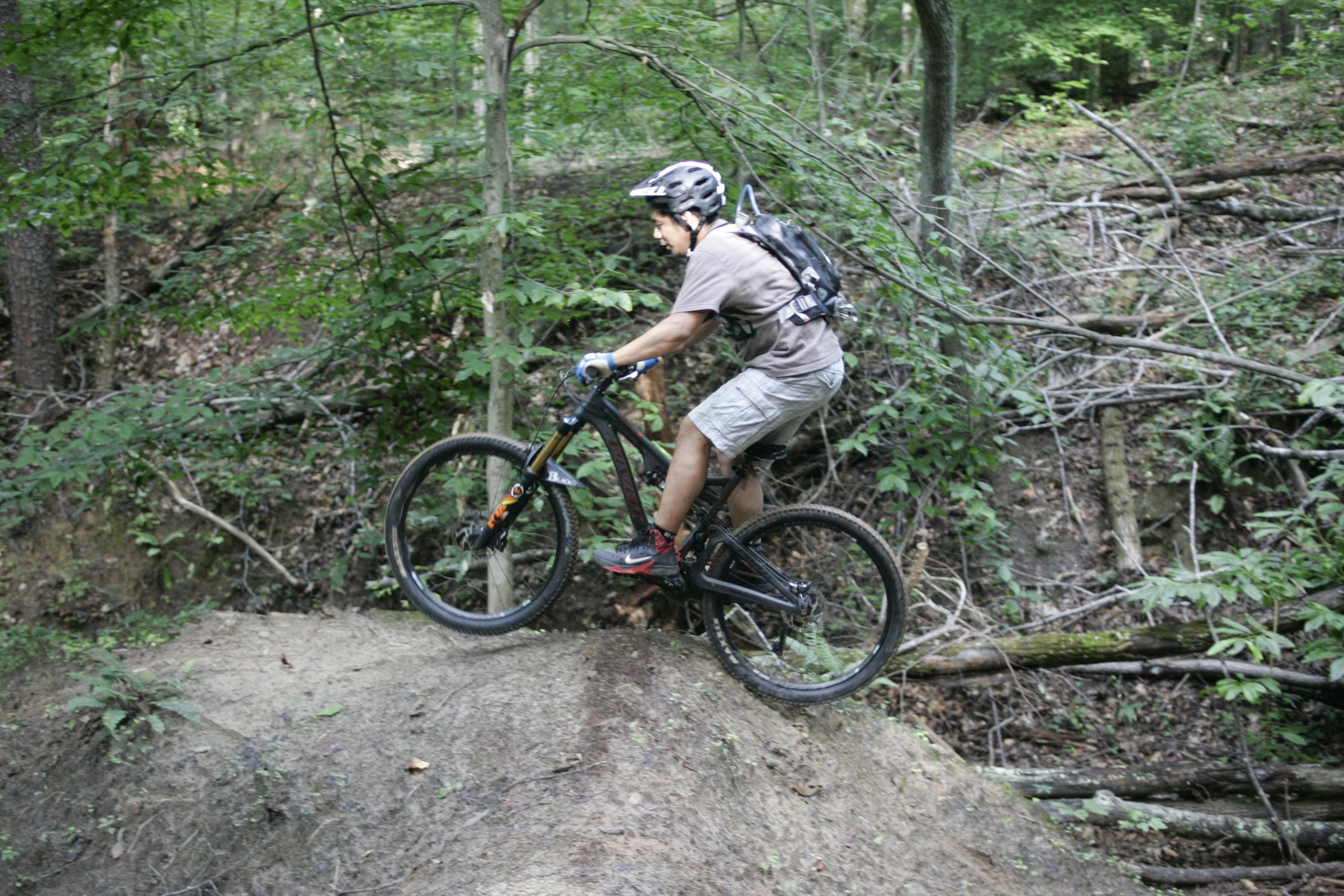 A mountain biker navigating a dirt trail, performing a wheelie on a steep slope in a lush green forest, surrounded by trees and fallen branches. Salem Lake mountain bike trail.