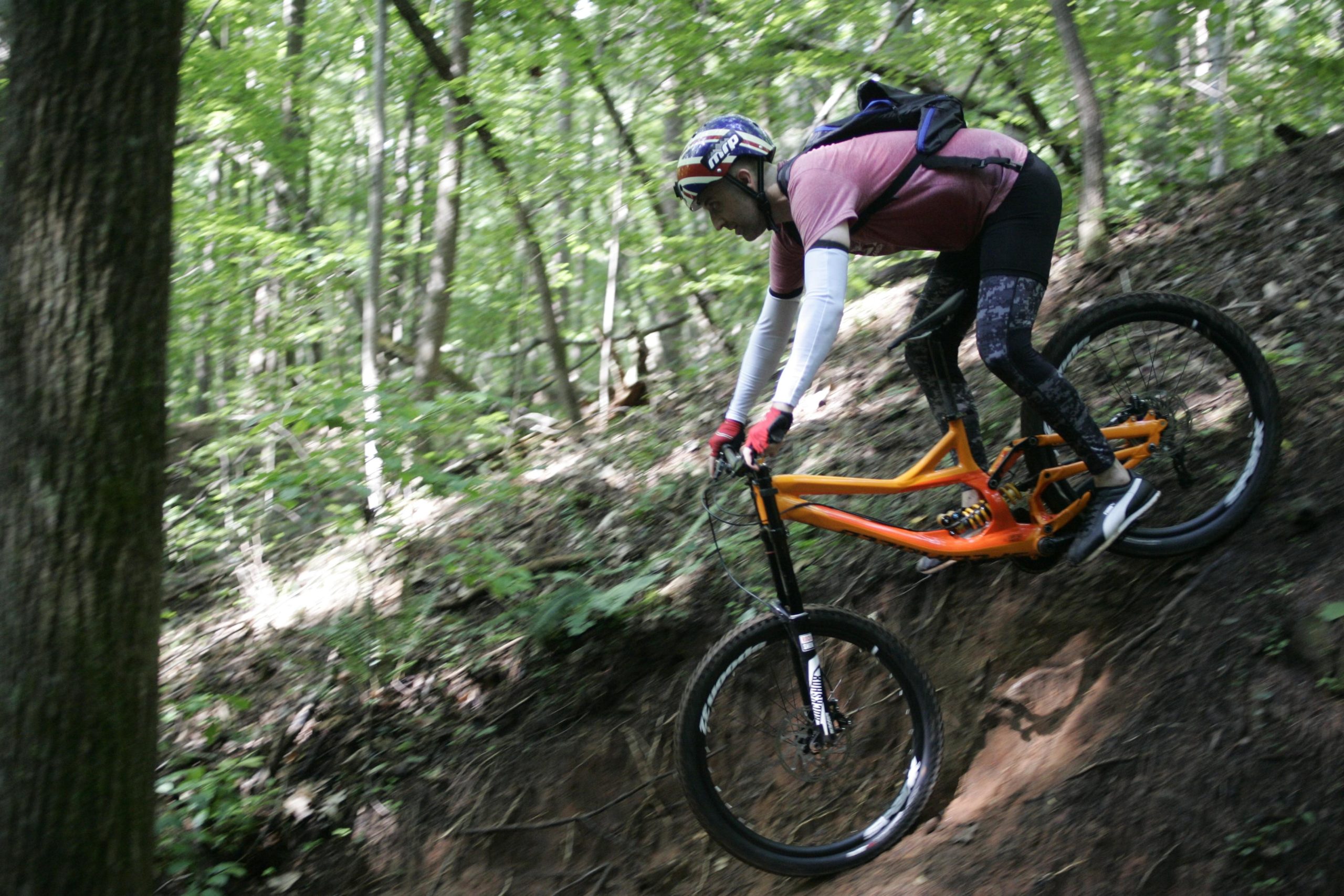 A mountain biker in a helmet and protective gear descends a steep, muddy slope in a wooded area, surrounded by lush green trees. The bike features a bright orange frame, and the rider is focused on navigating the terrain. Salem Lake mountain bike trail.