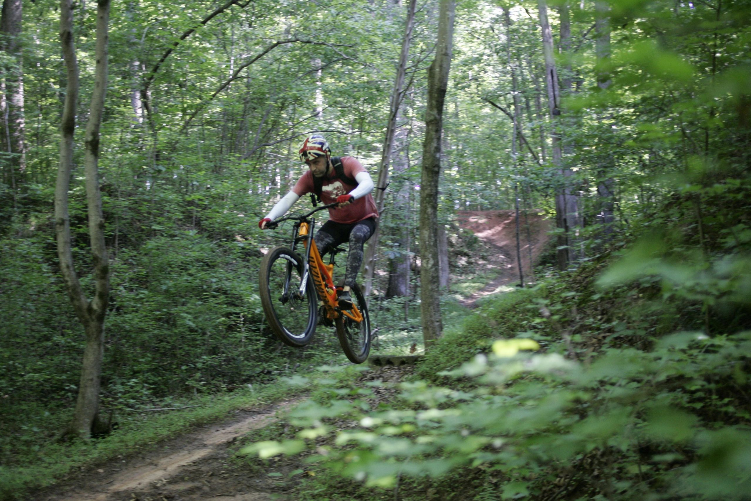 A mountain biker performing a jump on a dirt trail surrounded by lush greenery and trees. The rider is wearing a helmet and protective gear, capturing an action-packed moment in a forested landscape. Salem Lake mountain bike trail.