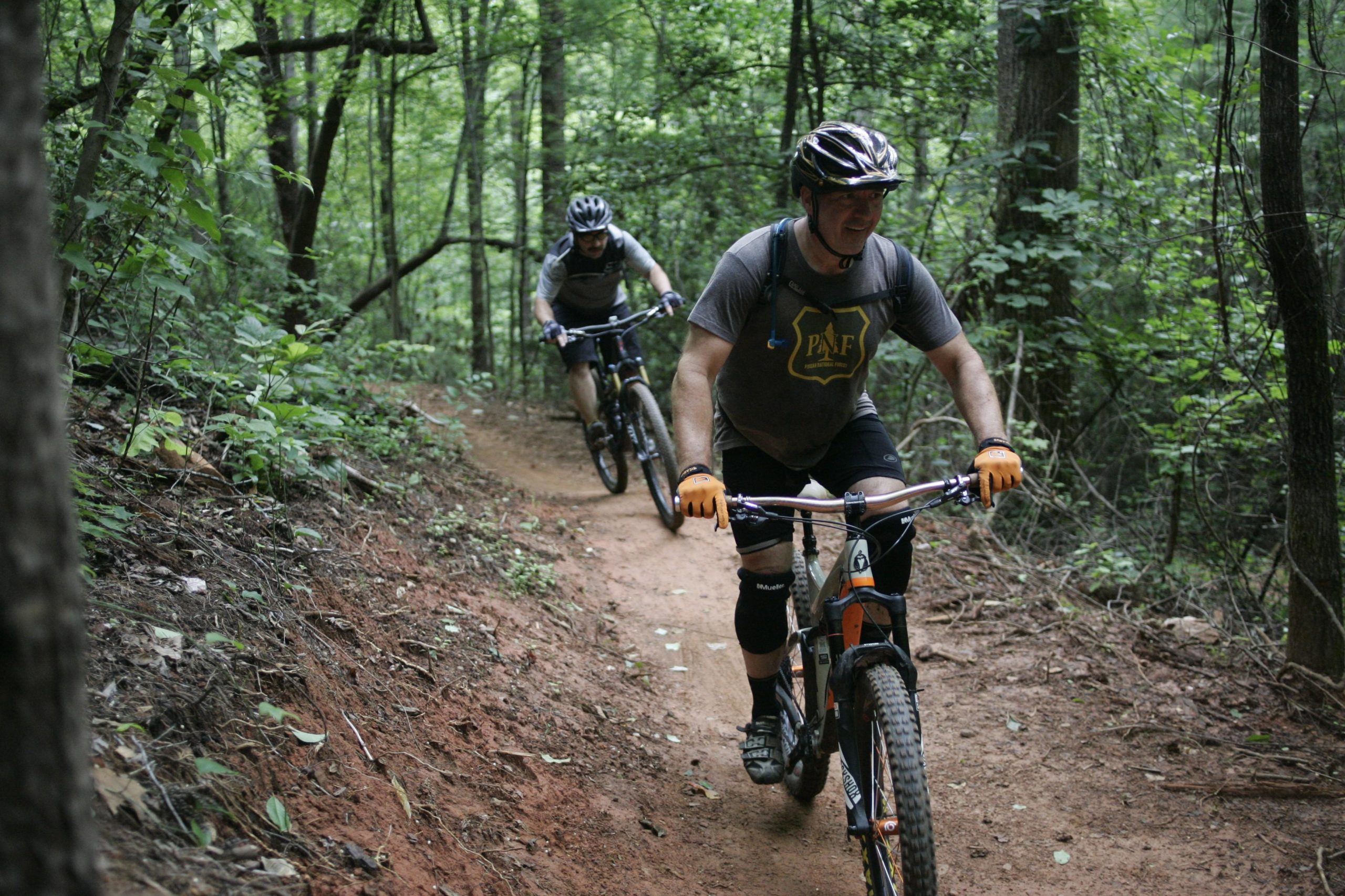 Two mountain bikers navigate a dirt trail surrounded by lush green trees. The path winds through a forest, with one biker in the foreground smiling as he rides. Both are wearing helmets and biking gear, enjoying their outdoor adventure in nature. Dark Mountain Trail mountain bike trail.